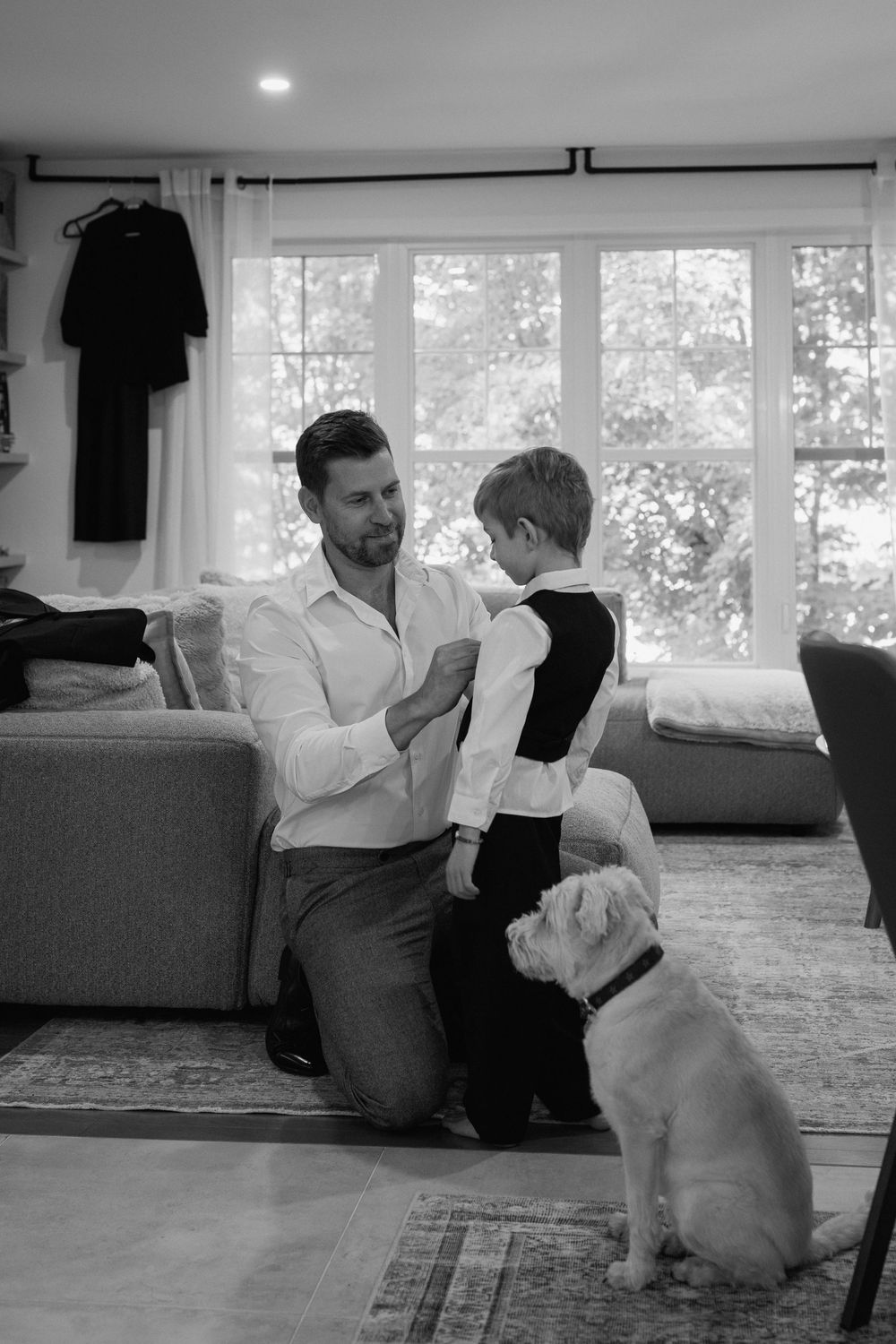 Black and white photo of a father kneeling to help his young son dress, with a bulldog watching in a bright living room.