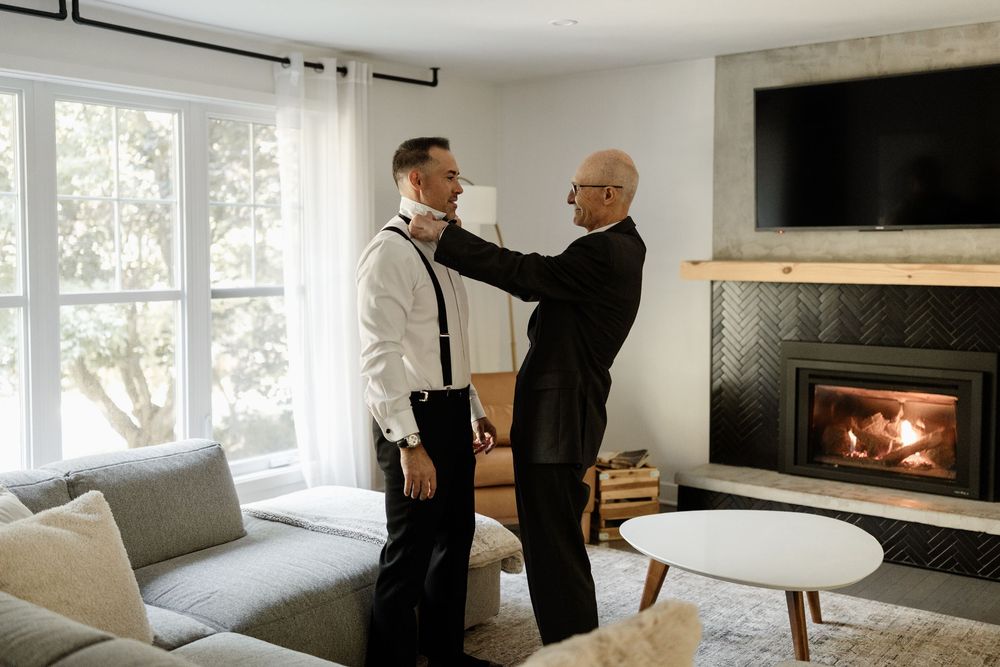 An older man adjusts a younger groom's bow tie in a cozy living room with a fireplace before a wedding.