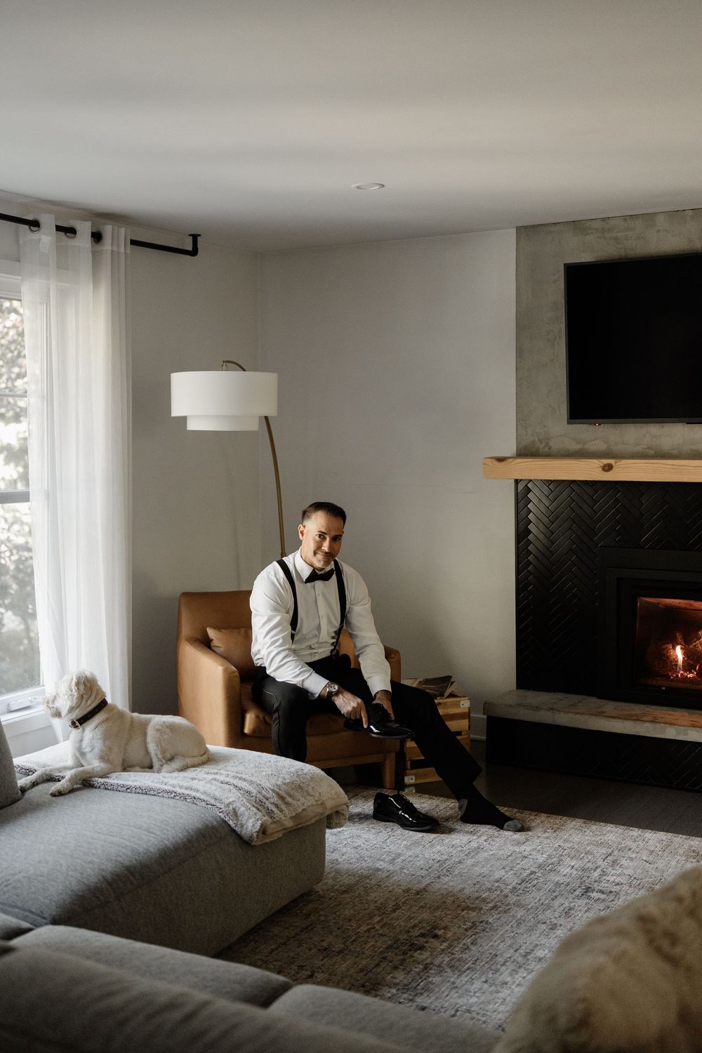 A groom in white shirt and suspenders sits by a fireplace putting on dress shoes, with a dog resting nearby.