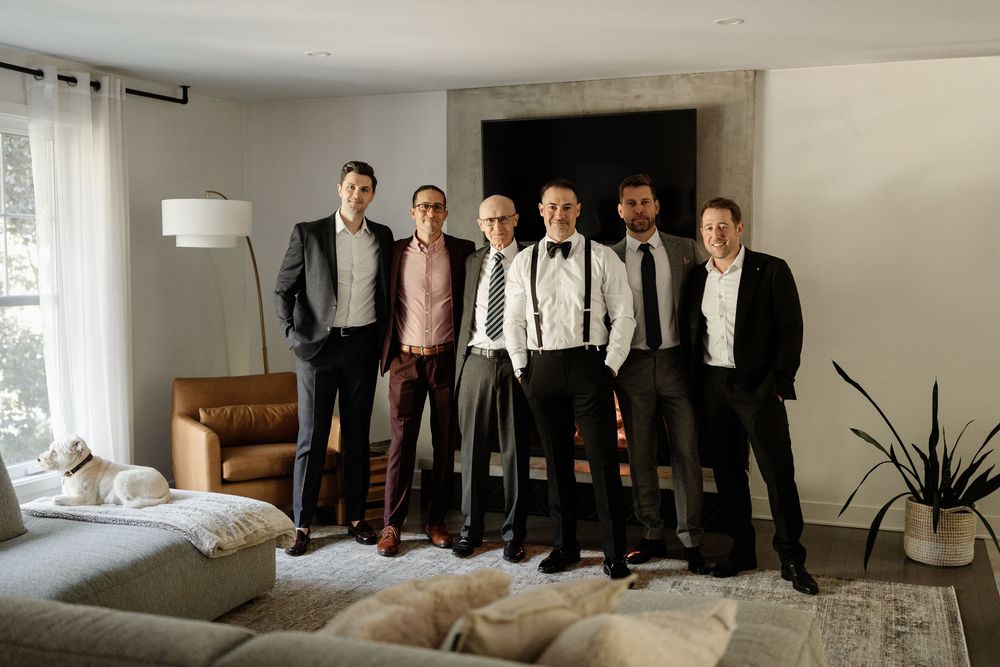 Six men in formal attire pose together smiling in a modern living room with neutral decor.