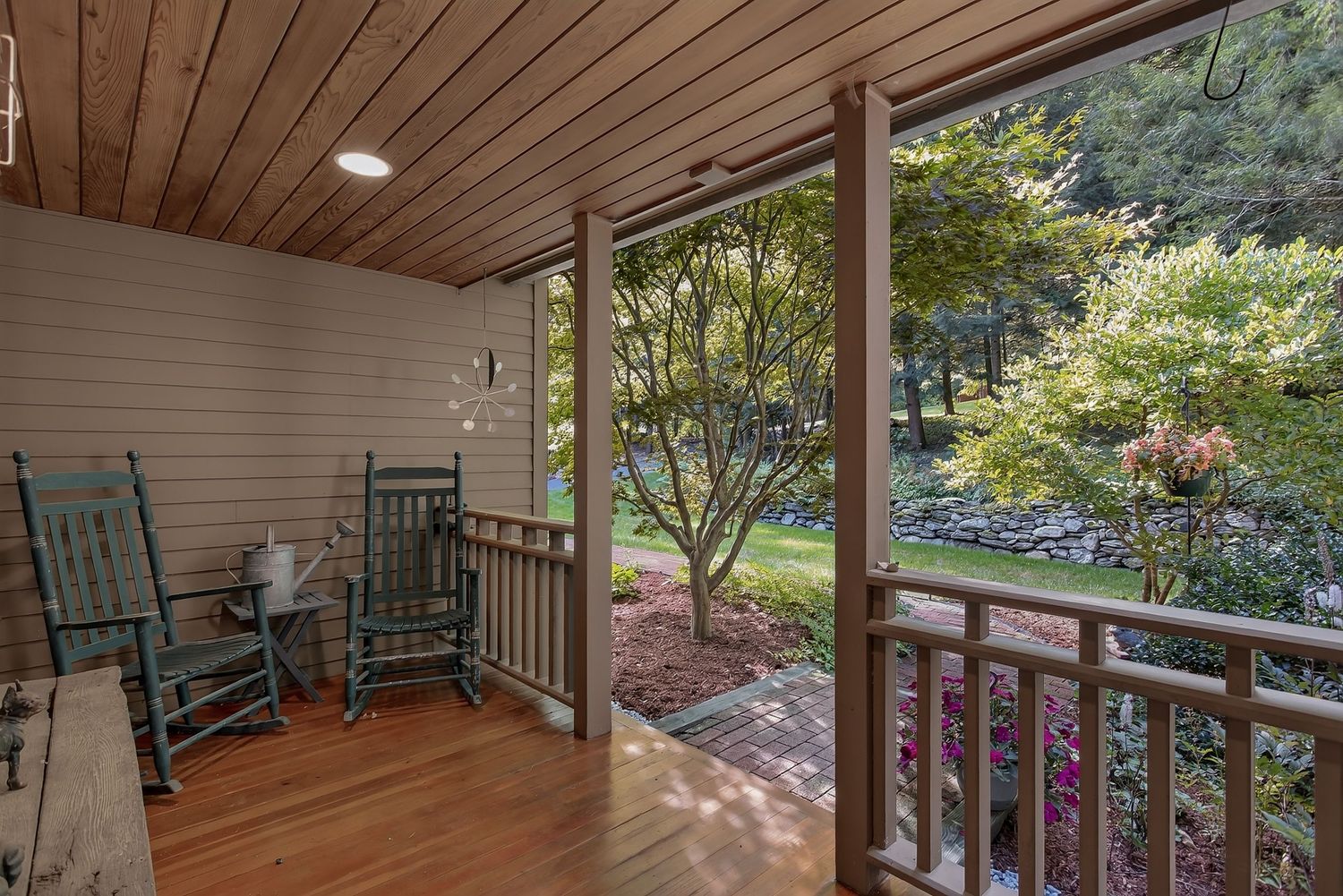 Covered wooden porch with rocking chairs overlooking a lush garden with colorful flowering shrubs.