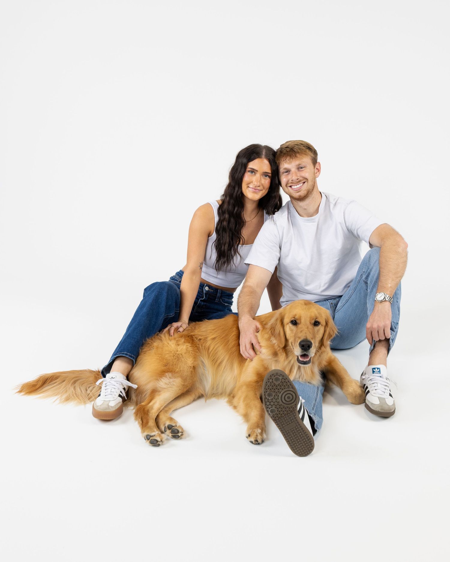 A smiling couple in white tees and jeans sit on a green velvet sofa with their golden retriever on a purple rug.