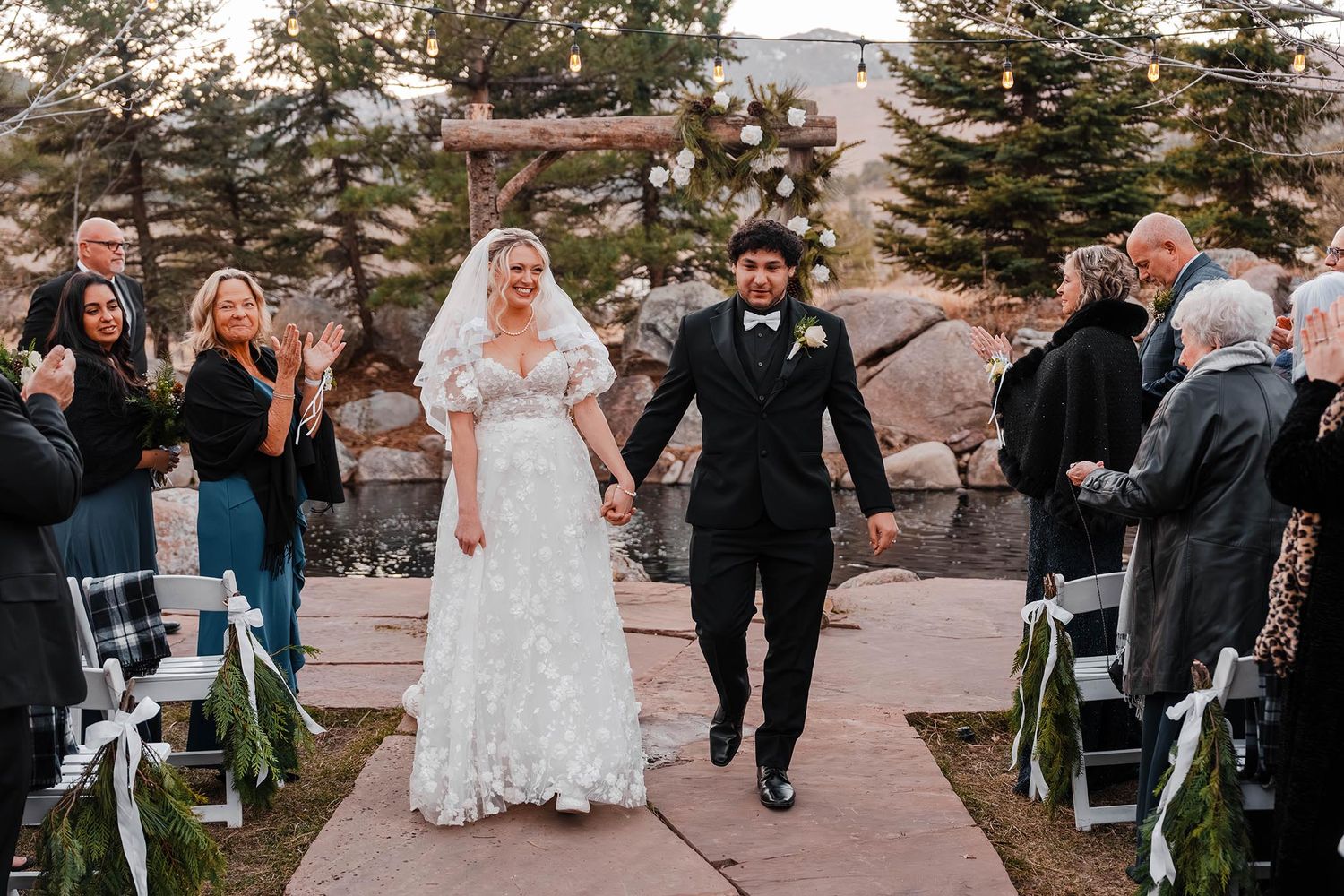 Bride and groom walk down the aisle after wedding ceremony outdoors, guests applauding, rocky landscape backdrop.