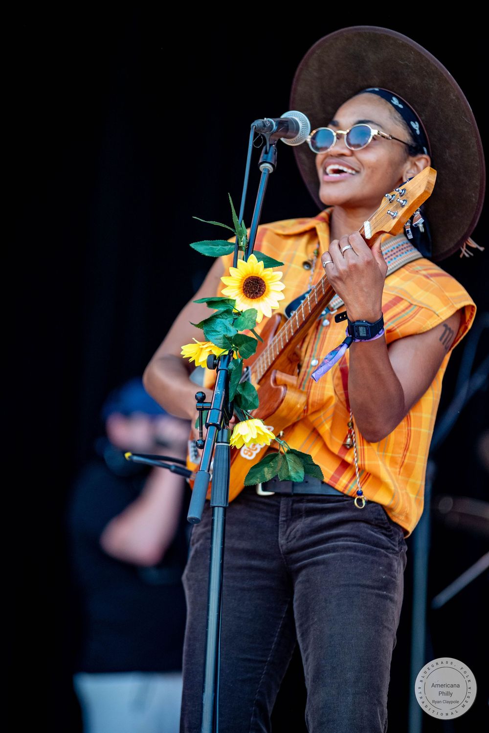A smiling musician in a wide-brim hat and sunglasses plays guitar on stage with sunflowers attached to the mic stand.