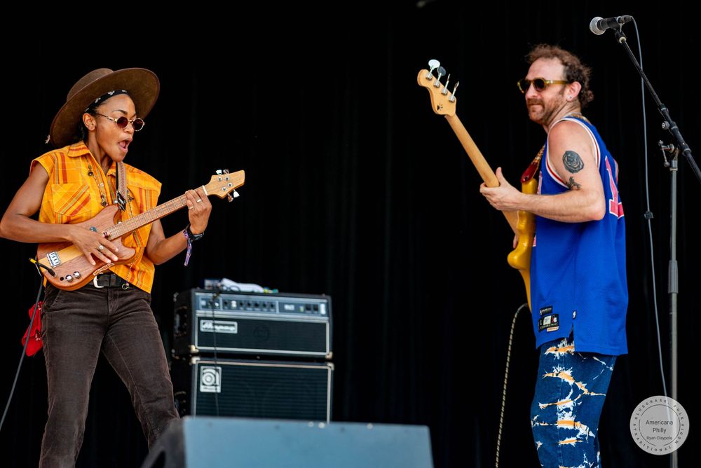 Two guitarists performing on outdoor stage, woman in yellow shirt and hat, man in blue sleeveless top with tattoos.