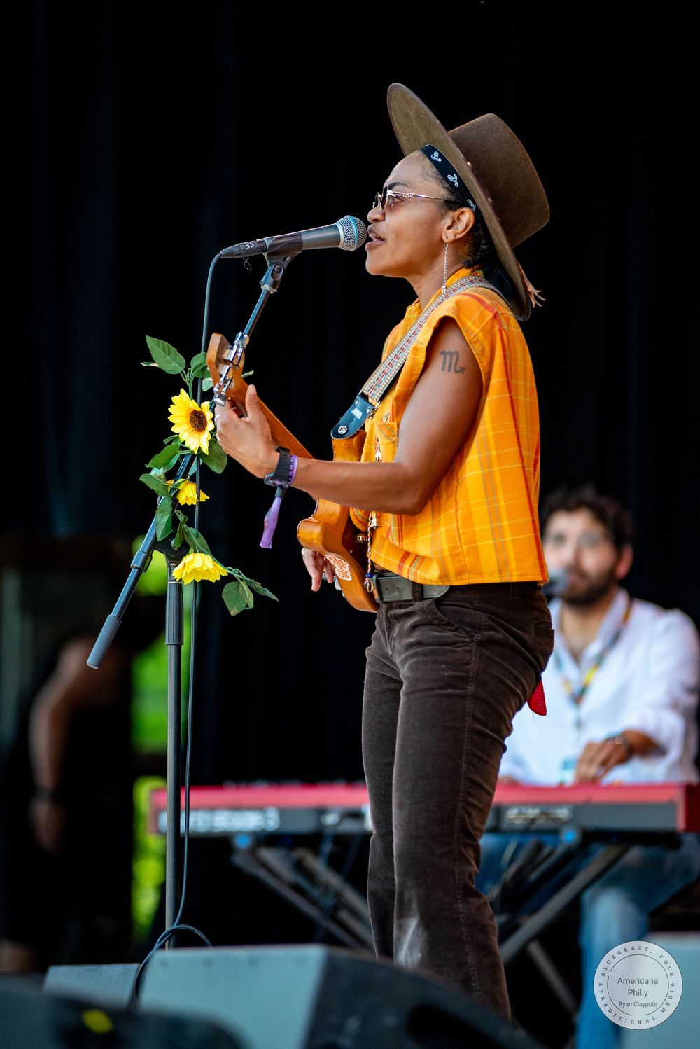 Musician in orange top and brown hat plays guitar at outdoor festival stage decorated with sunflowers.