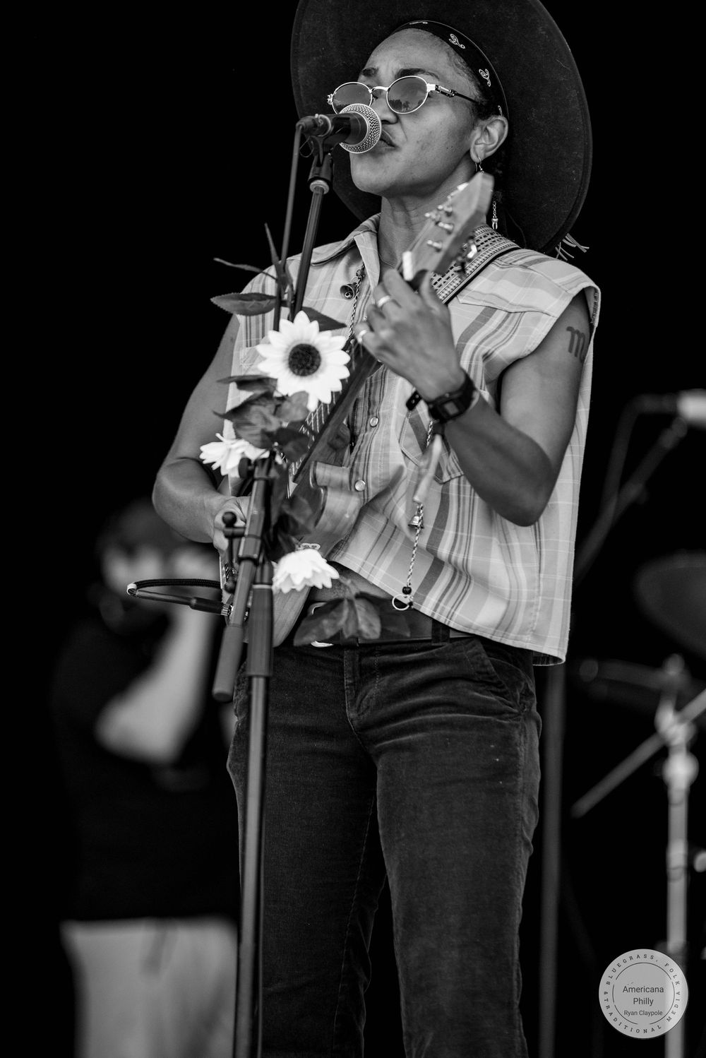 Black and white photo of a performer singing at a mic, holding flowers, wearing a wide-brim hat and sunglasses on stage.