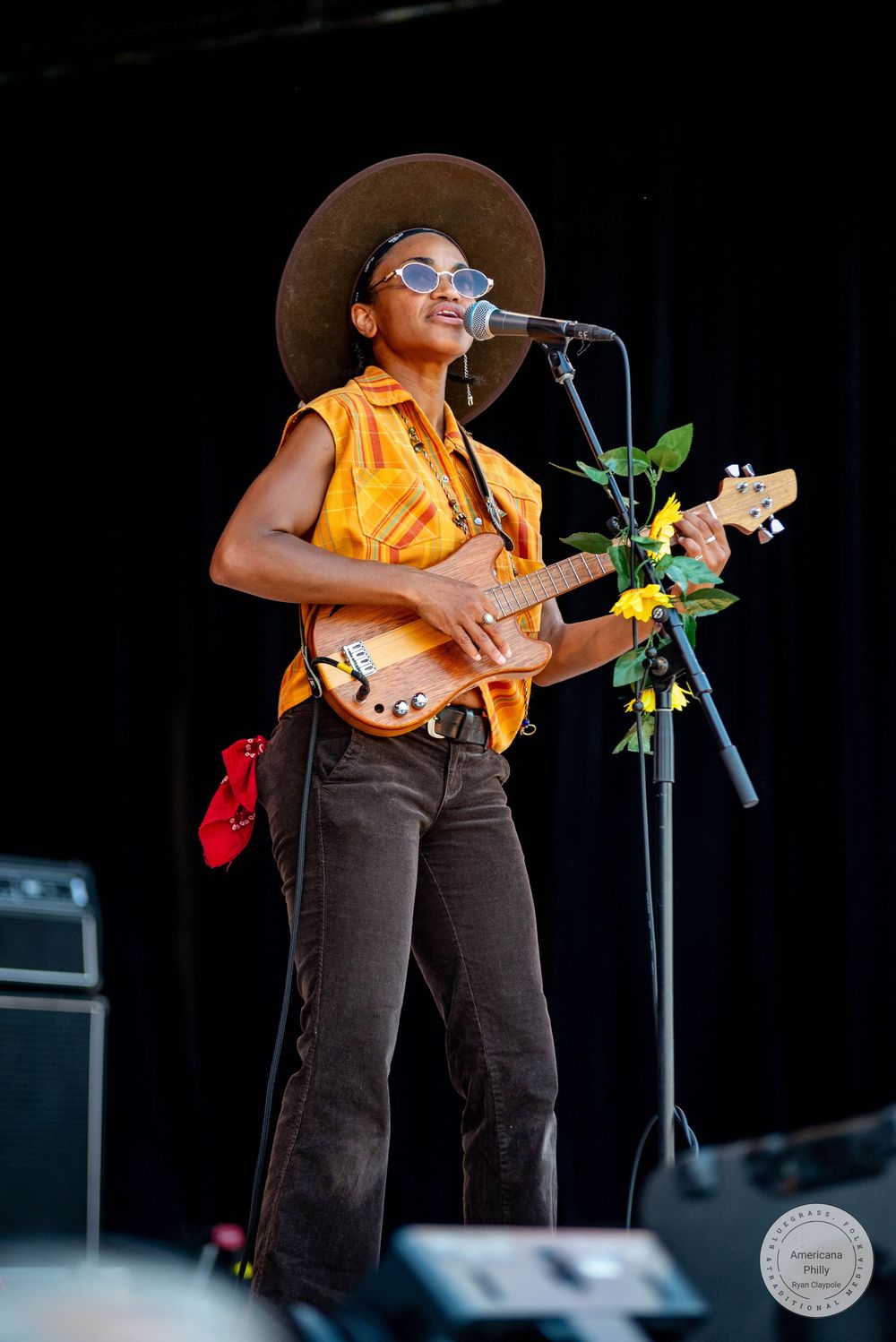 A woman in a wide-brim hat and orange vest plays guitar on stage with yellow flowers on the neck.
