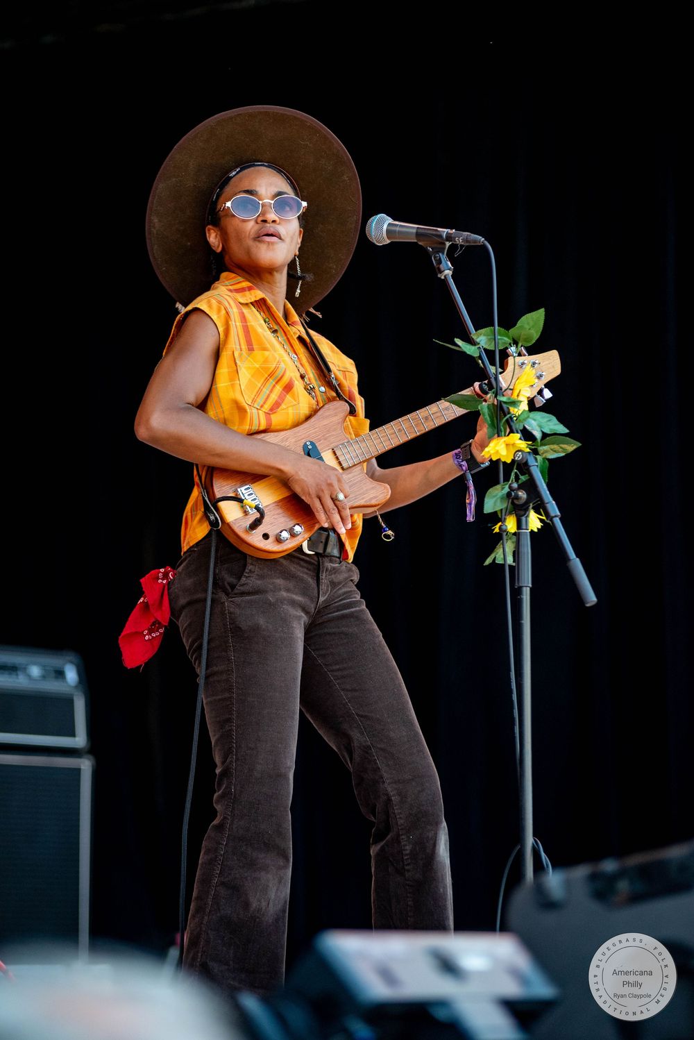A woman in a wide-brim hat and sunglasses plays electric guitar on stage, wearing an orange vest and dark flared pants.