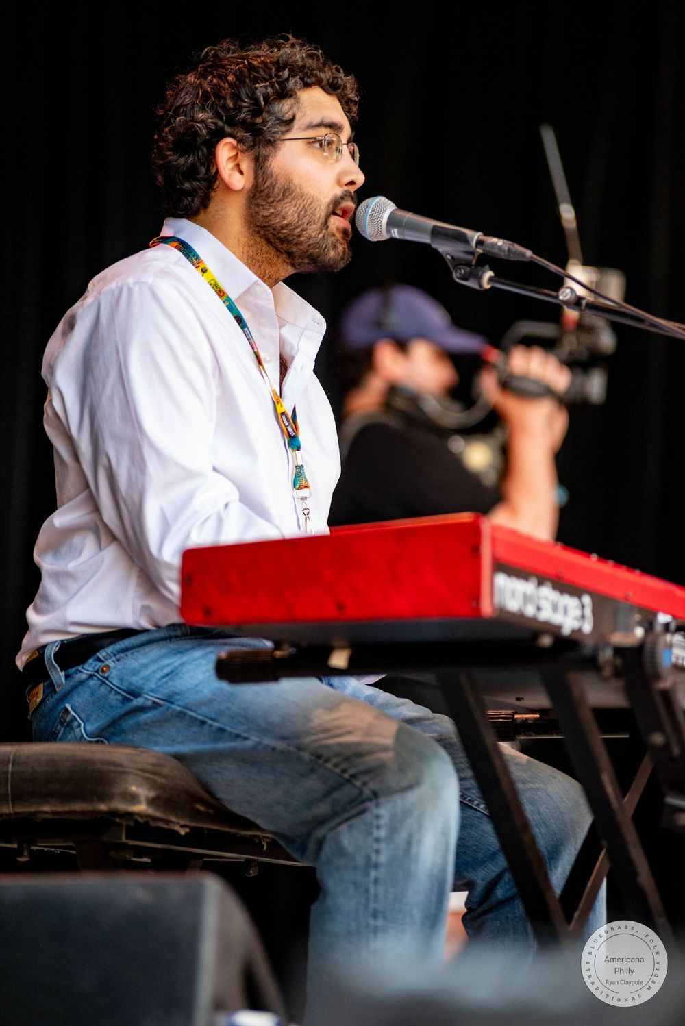Bearded musician playing a red keyboard synthesizer on stage while singing into a microphone at an outdoor concert.