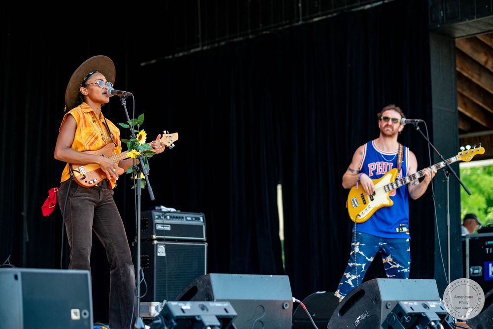 Two musicians perform on outdoor stage, woman playing ukulele and man playing guitar at a music festival.
