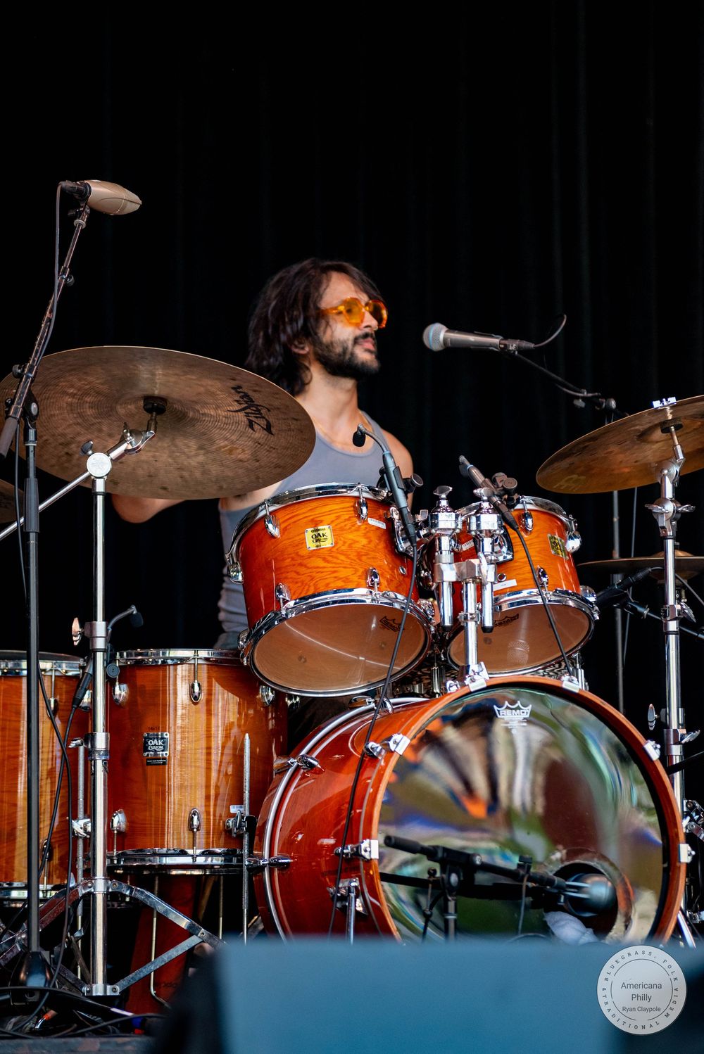 Male drummer with dreadlocks and orange sunglasses playing an orange drum kit on an outdoor stage.