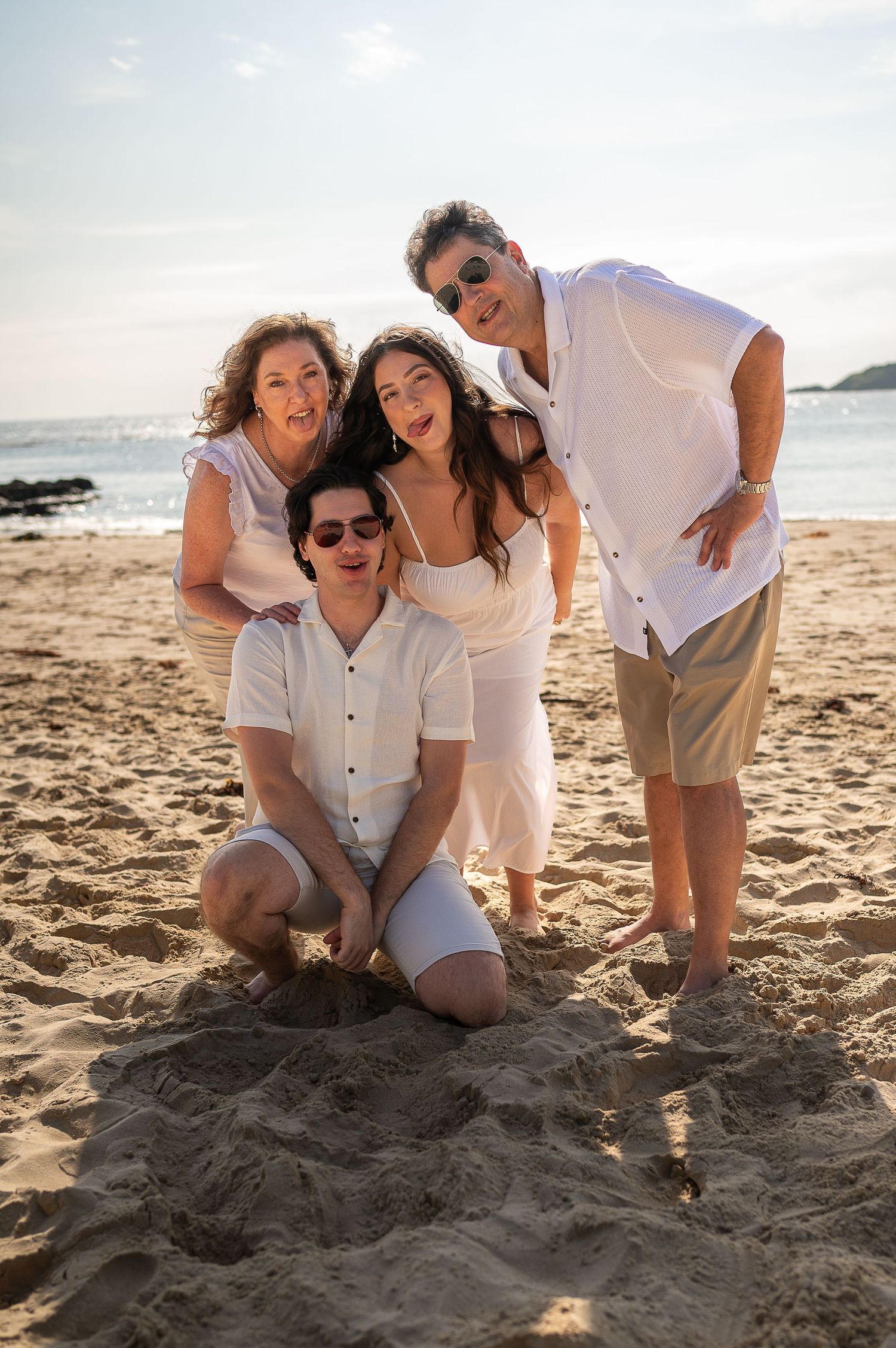Family of four posing together on a sandy beach at sunset, all wearing white outfits, smiling joyfully.