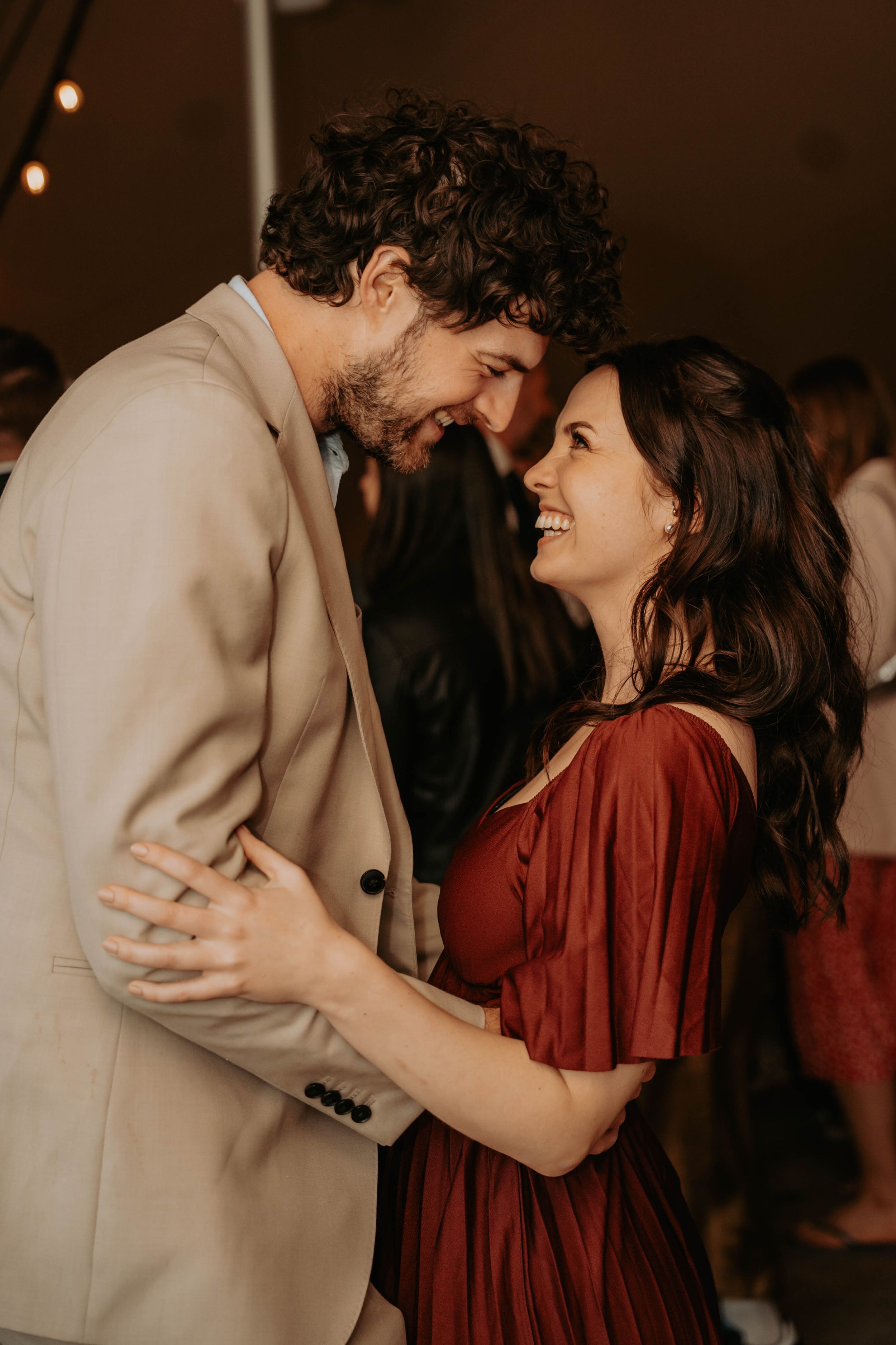 A couple shares a joyful dance, the woman in a red dress smiling as they embrace at a warm, festive event.