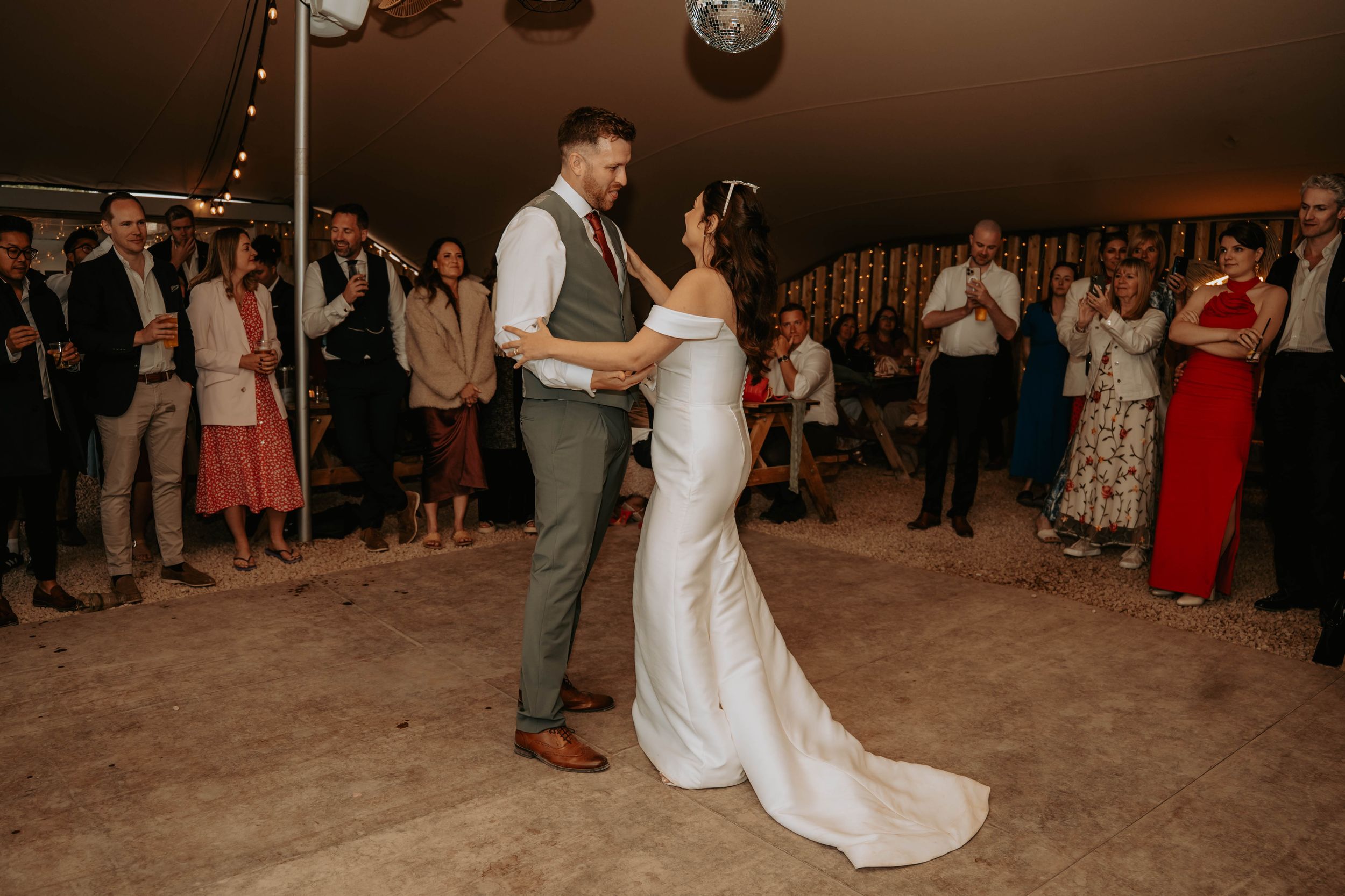 Bride and groom share their first dance in a elegant marquee tent surrounded by applauding wedding guests.
