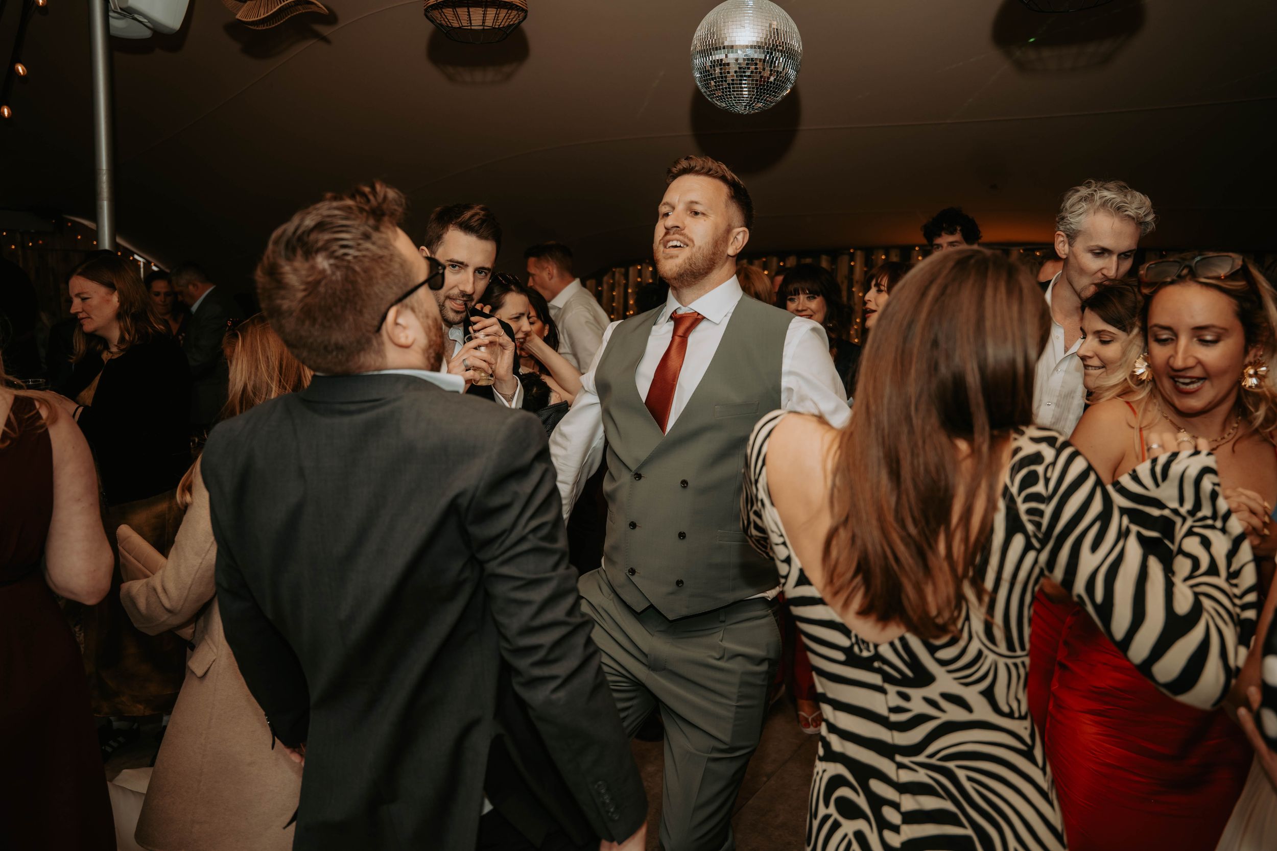 Groom in grey suit dancing with guests under disco ball at lively wedding reception party.
