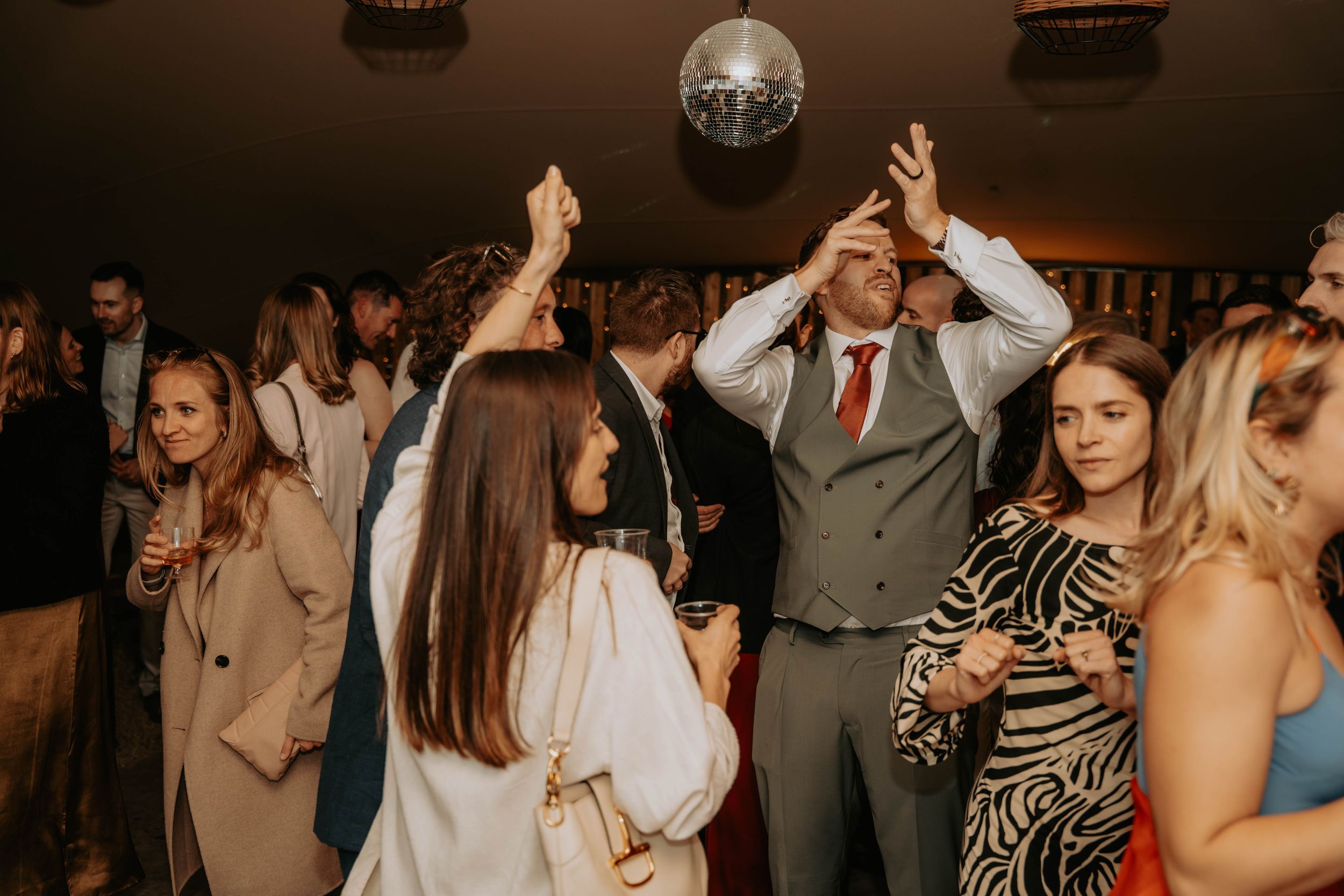 Guests dancing at a wedding reception under a disco ball in a warmly lit venue.