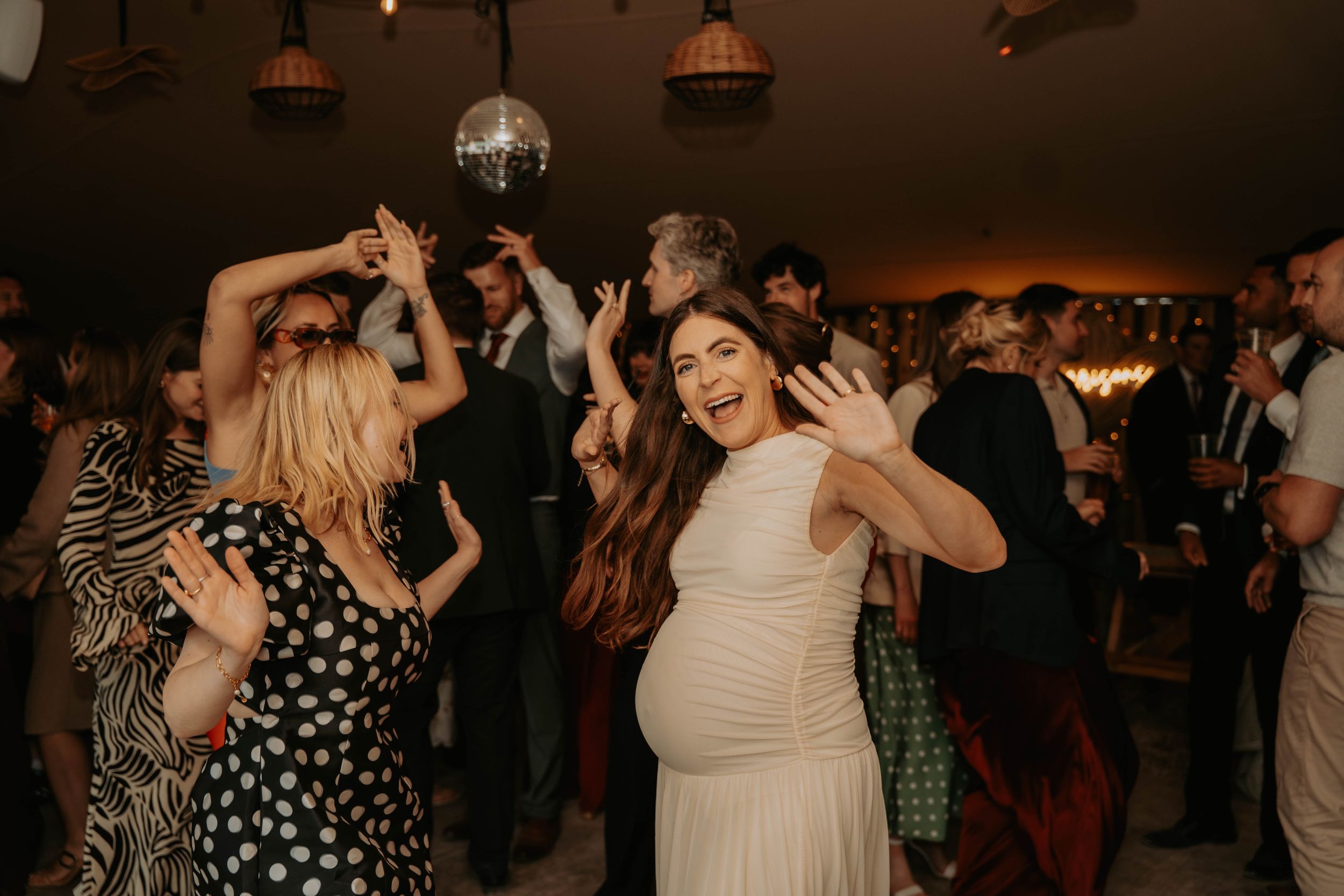 Pregnant bride in white dress dancing joyfully at wedding reception with guests and disco ball.