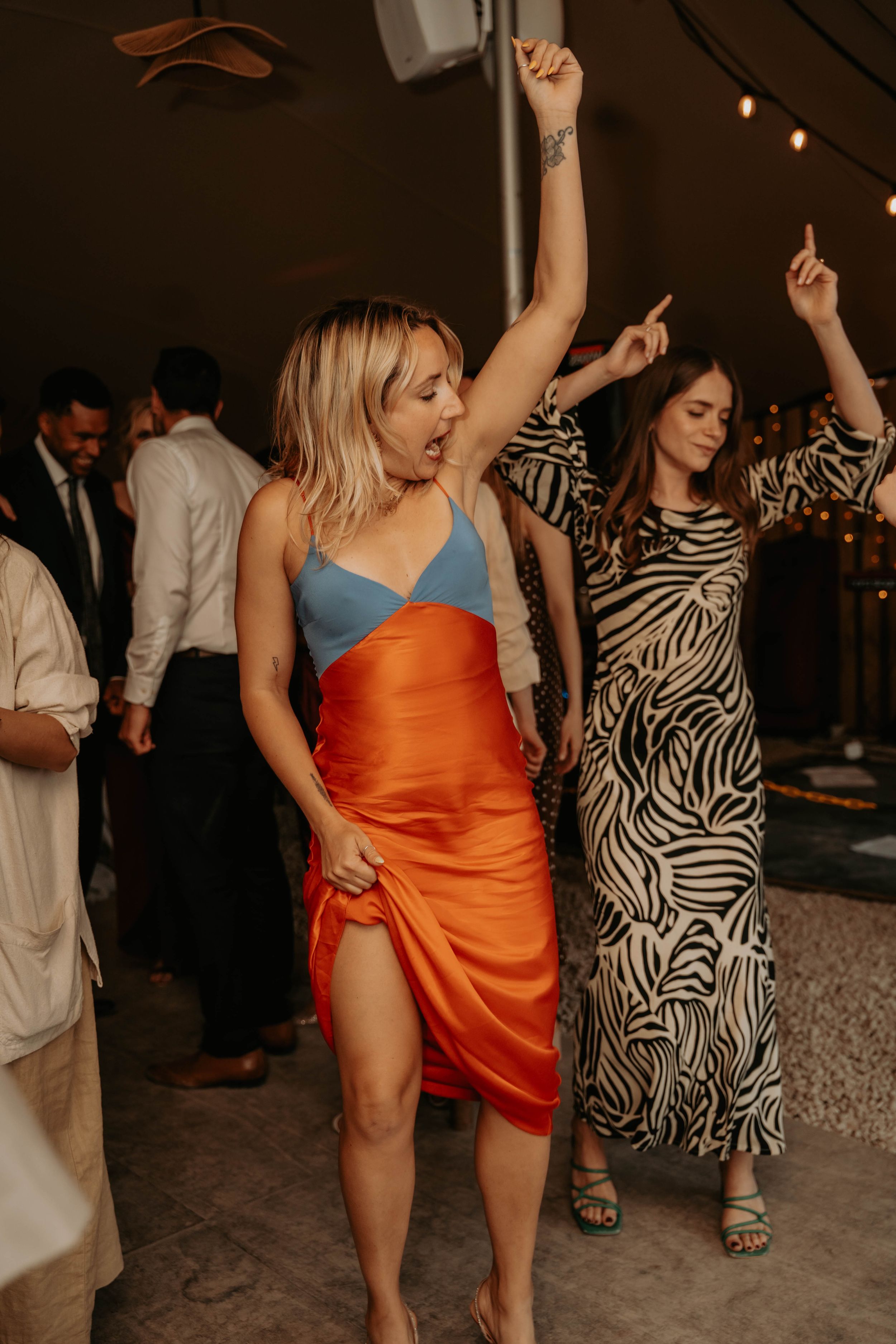 Two women dancing joyfully at a party, one in an orange and blue dress, the other in a zebra print gown.