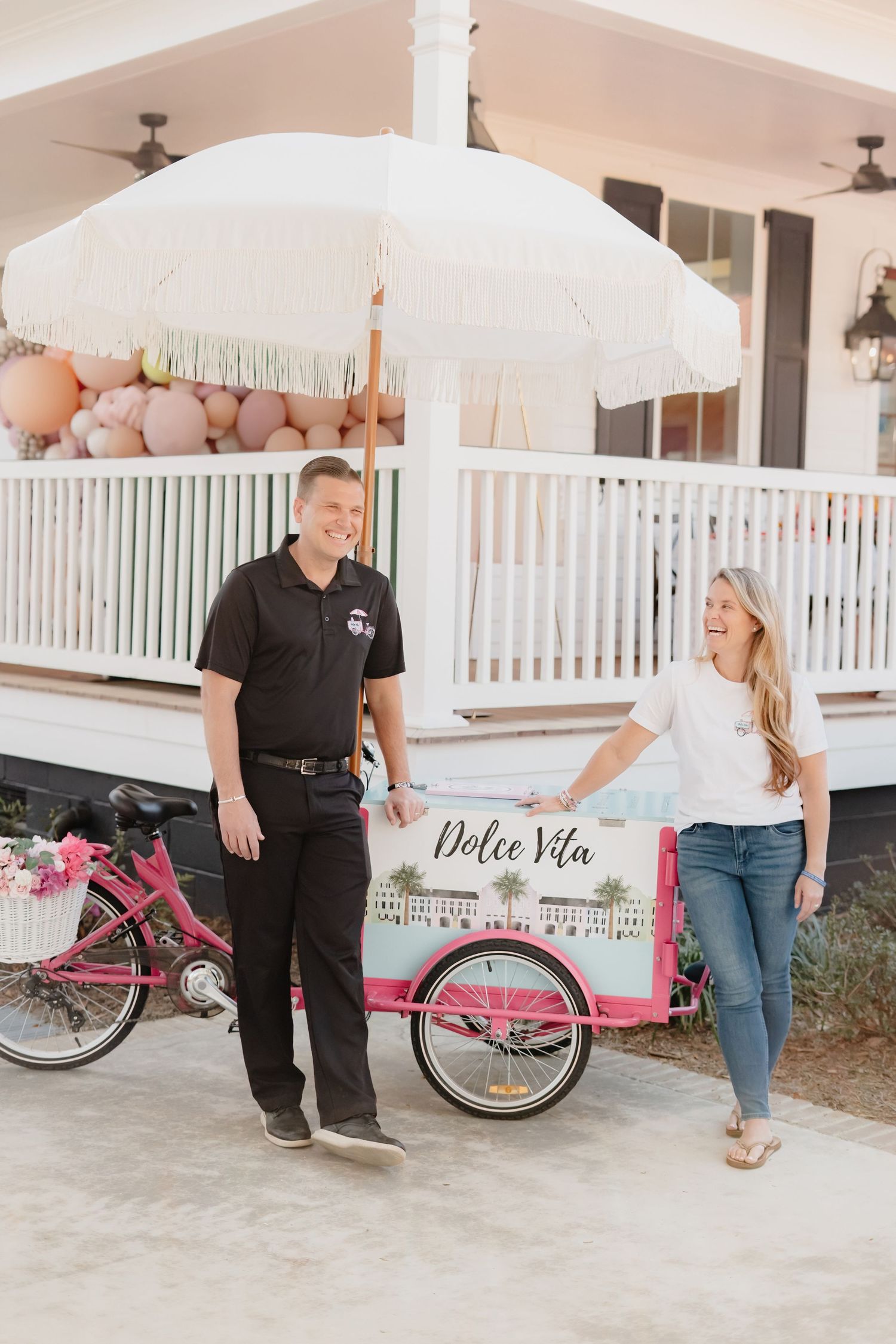 A man and woman smile beside a pink Dolce Vita ice cream cart with a white umbrella outside a charming venue.