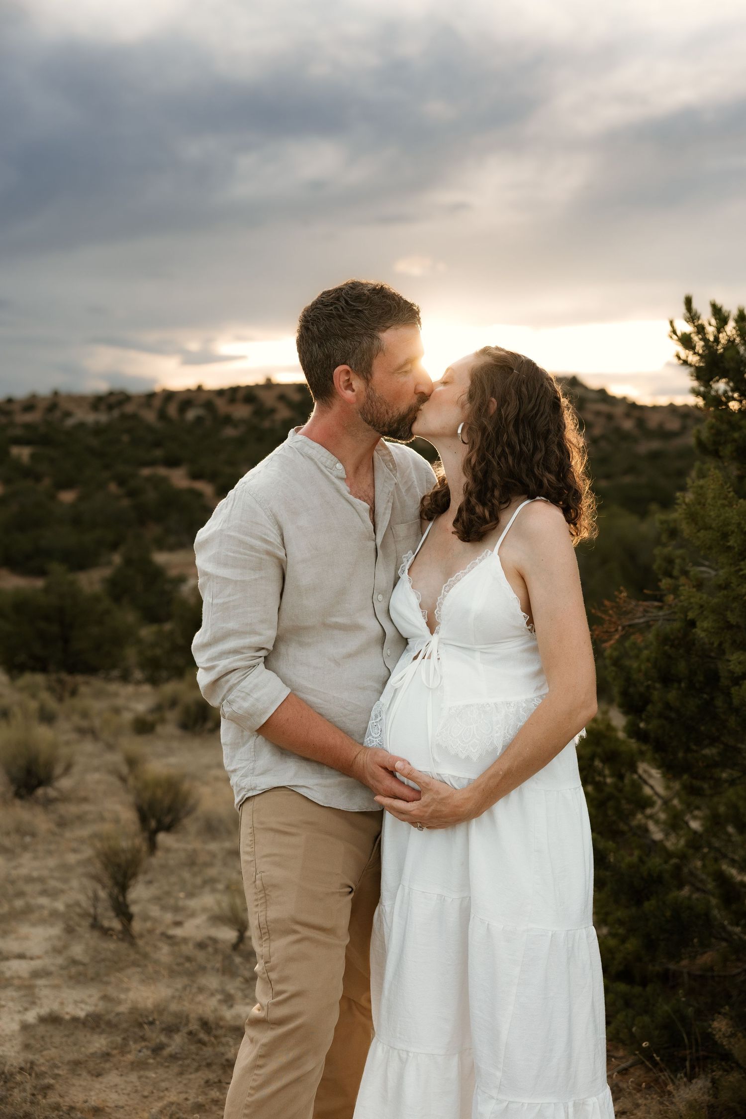 Couple sharing a kiss at sunset in a desert landscape, pregnant woman in white dress with partner in linen shirt.