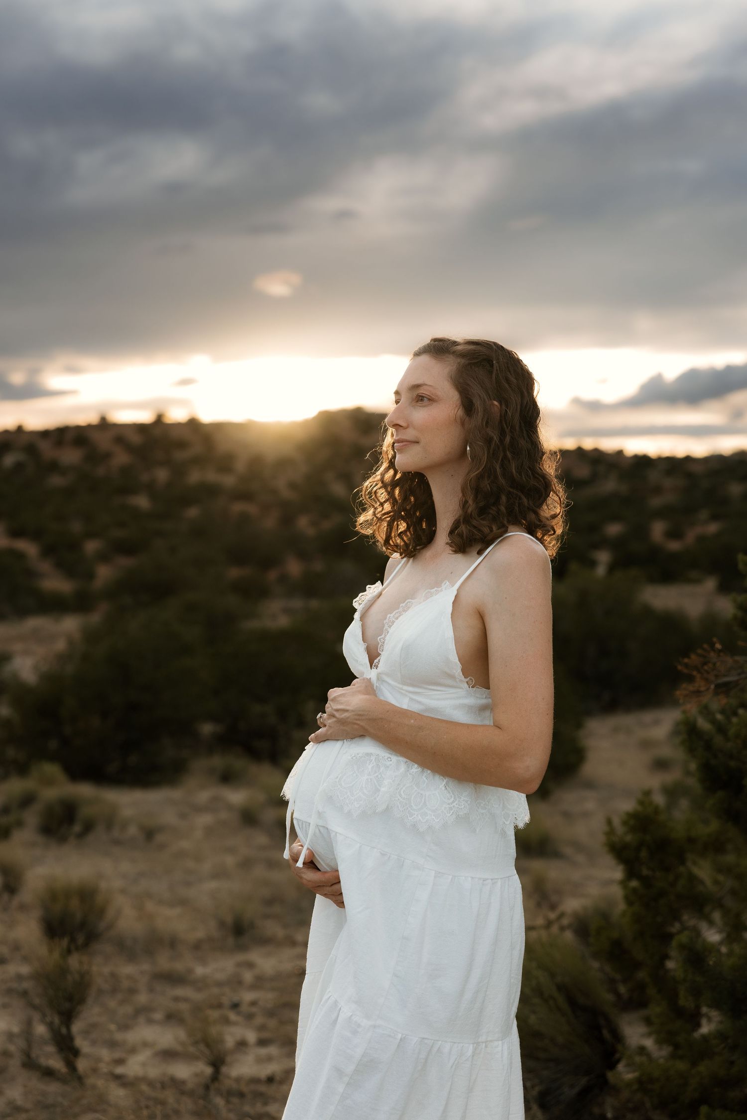 Pregnant woman in white dress holding belly at sunset in desert landscape.