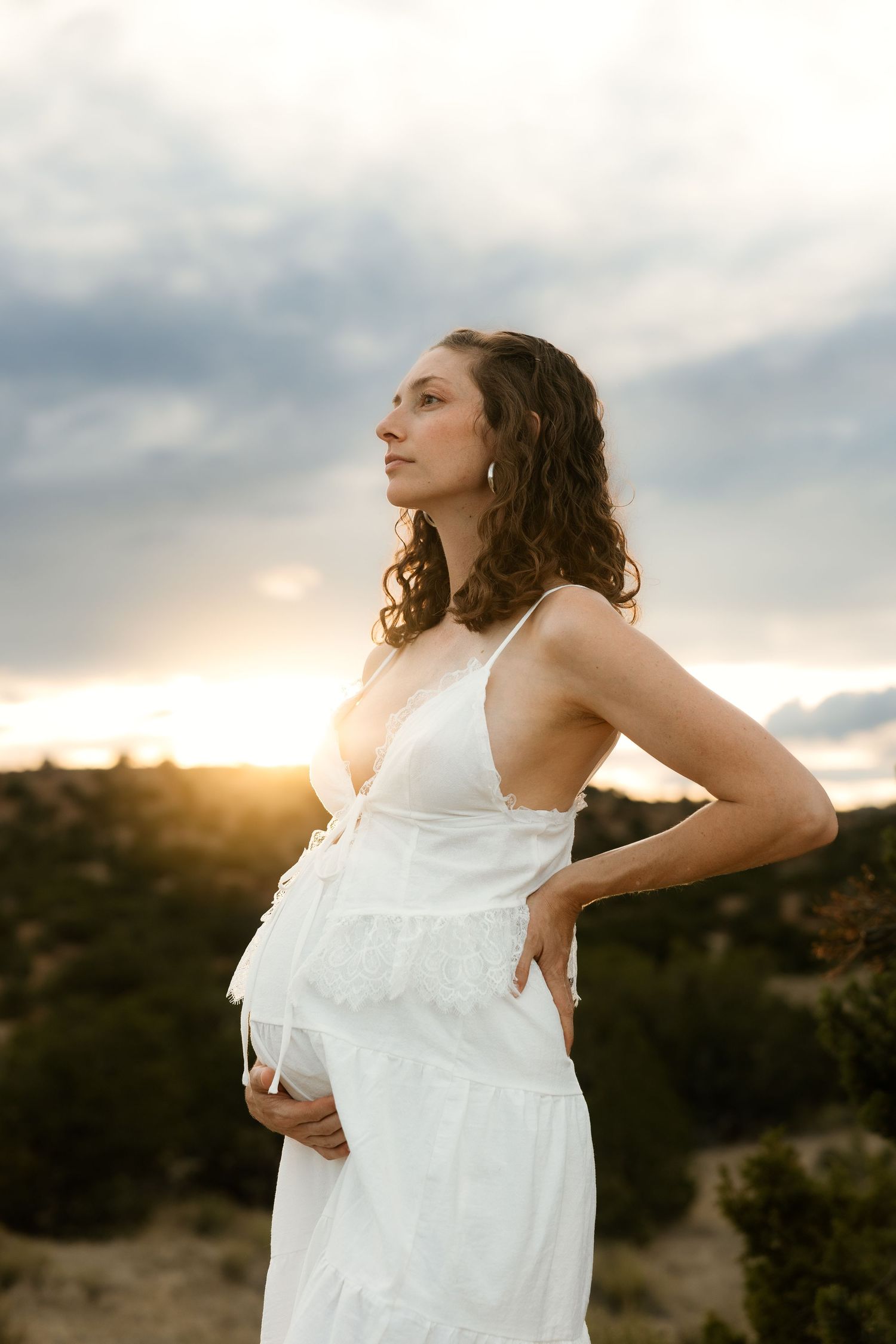 Pregnant woman in white dress holding belly at sunset outdoors.