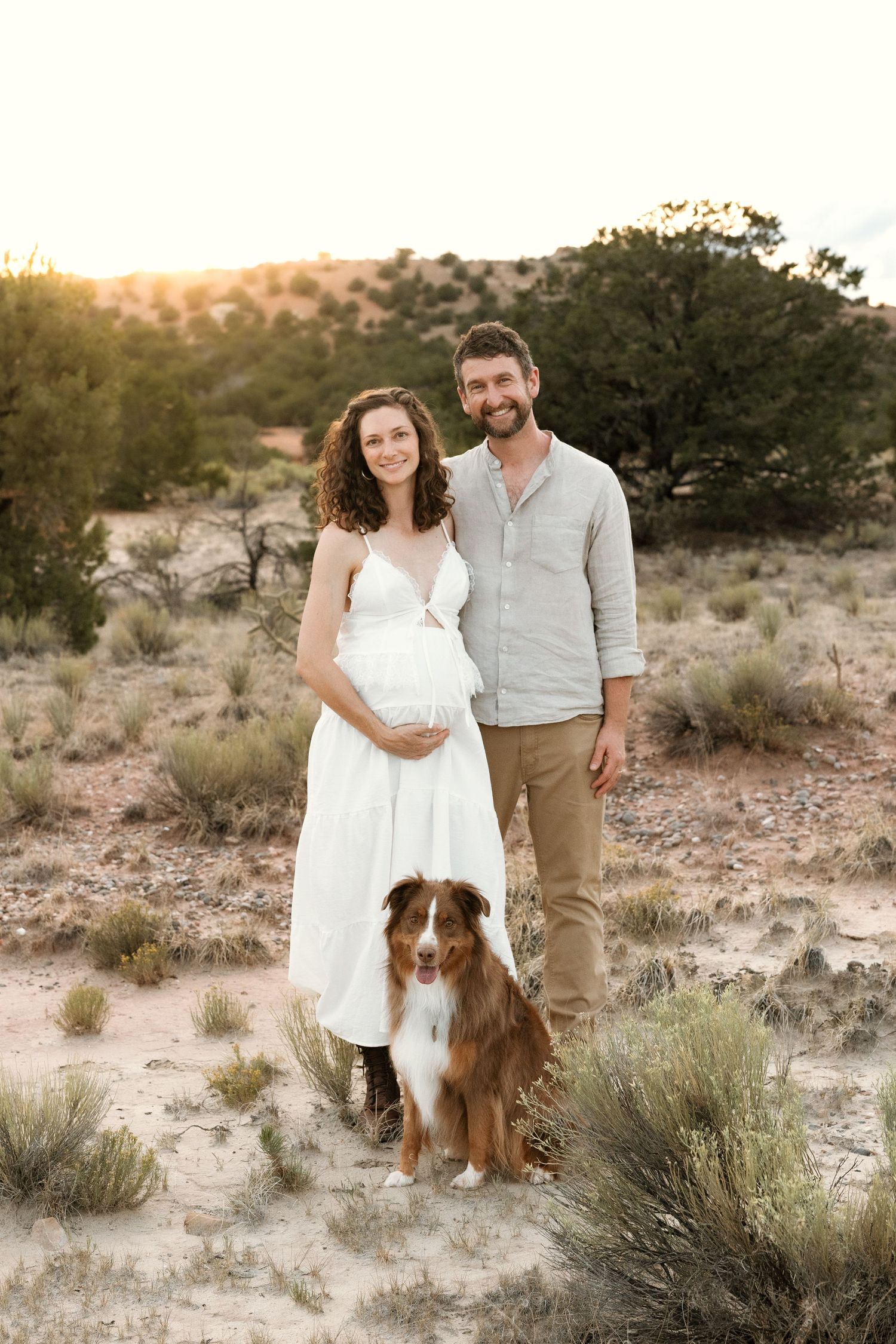 Pregnant couple poses with Australian Shepherd dog in desert landscape at golden hour sunset.
