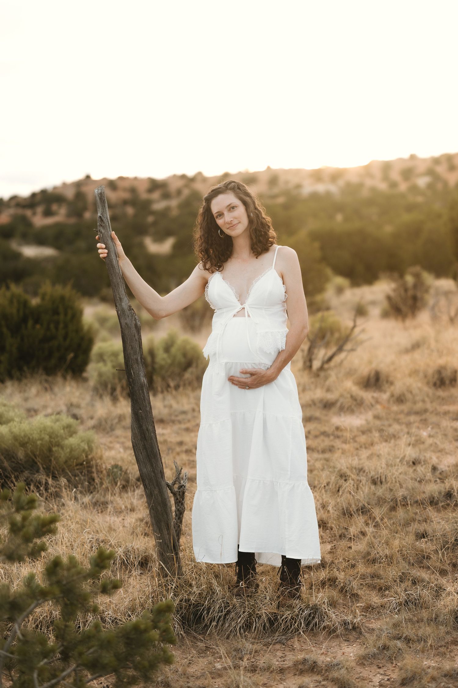 Pregnant woman in white dress holding belly outdoors at golden hour in a desert landscape.