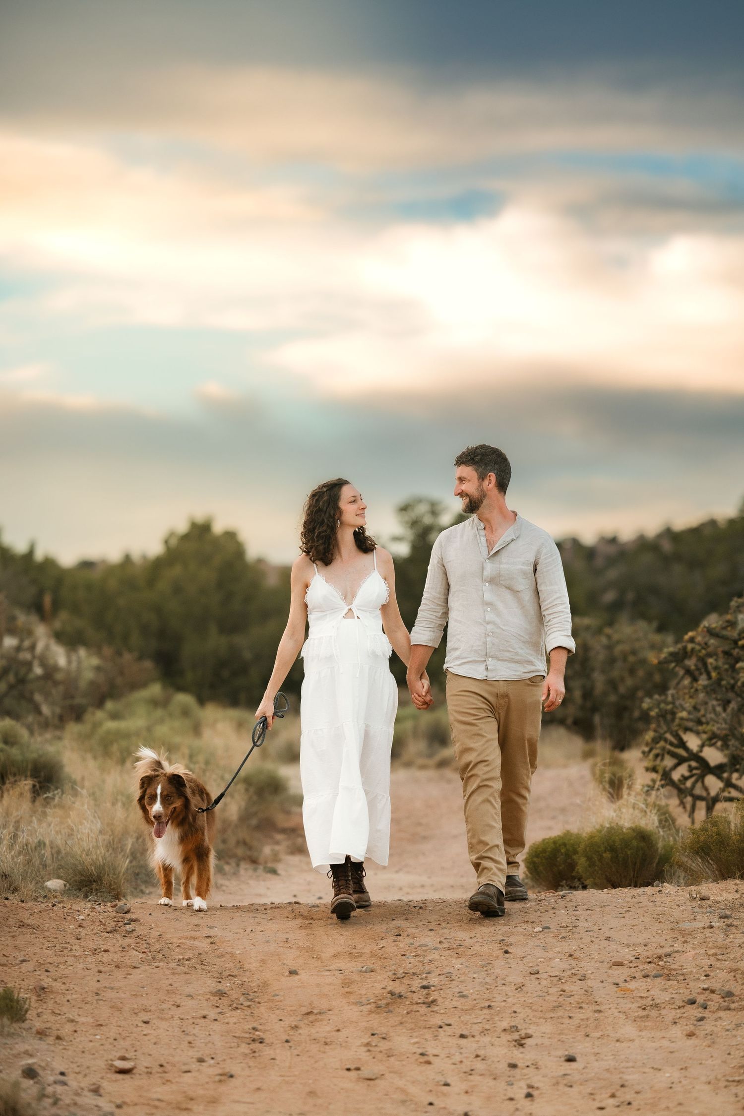 Couple walking dog on dirt path at sunset, woman in white dress, man in casual attire, desert landscape background.