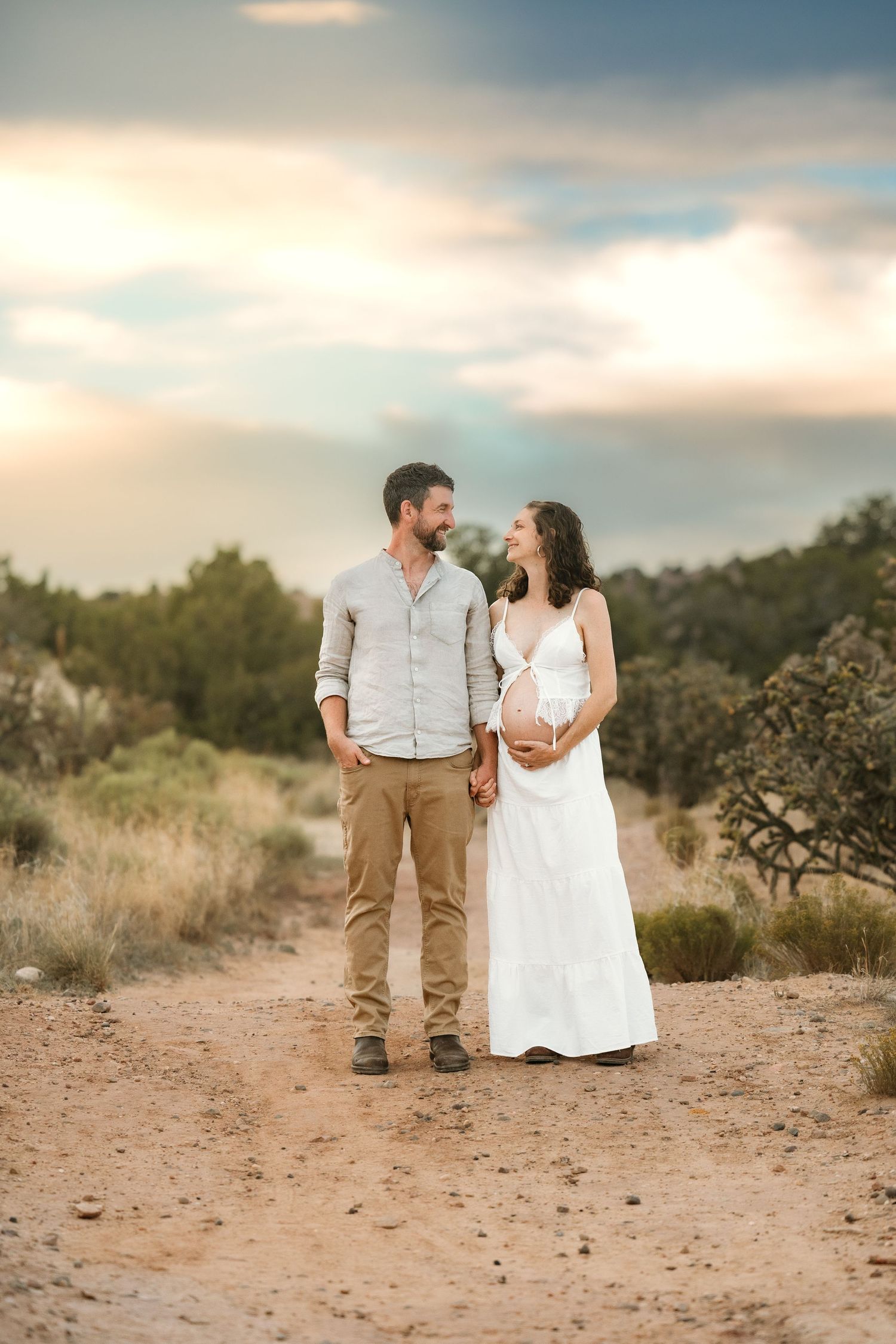 Couple holding hands on desert path, pregnant woman in white dress, man in casual attire, sunset sky backdrop.