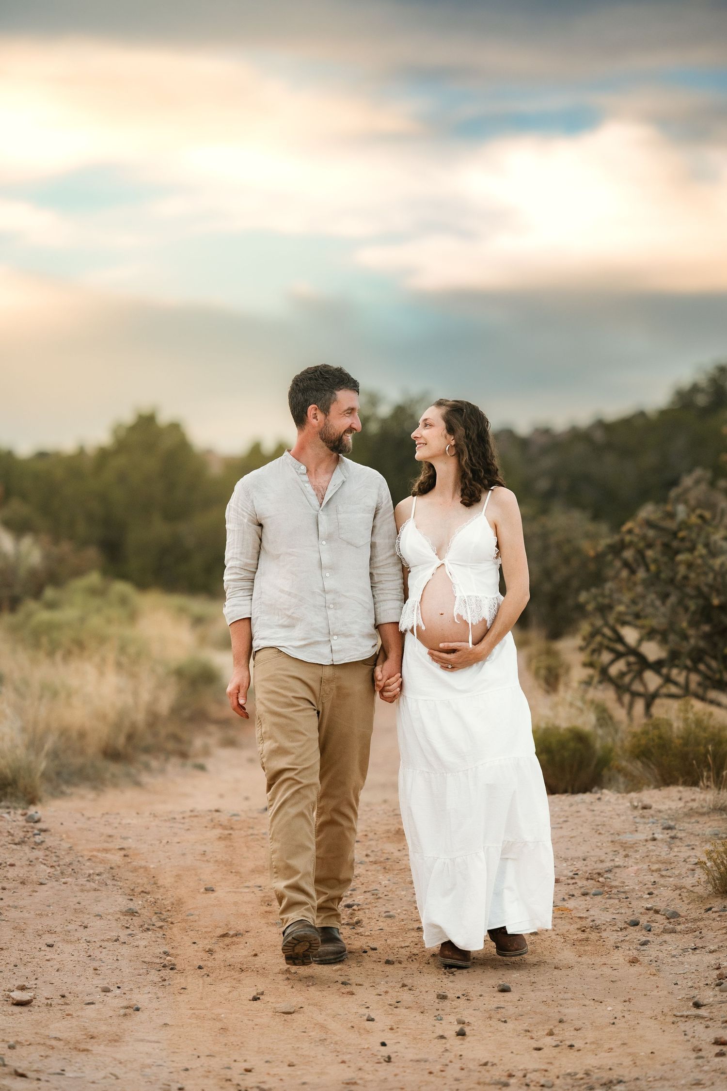 Expecting couple walking hand-in-hand on a dirt path at sunset, woman in white maternity dress smiling at partner.