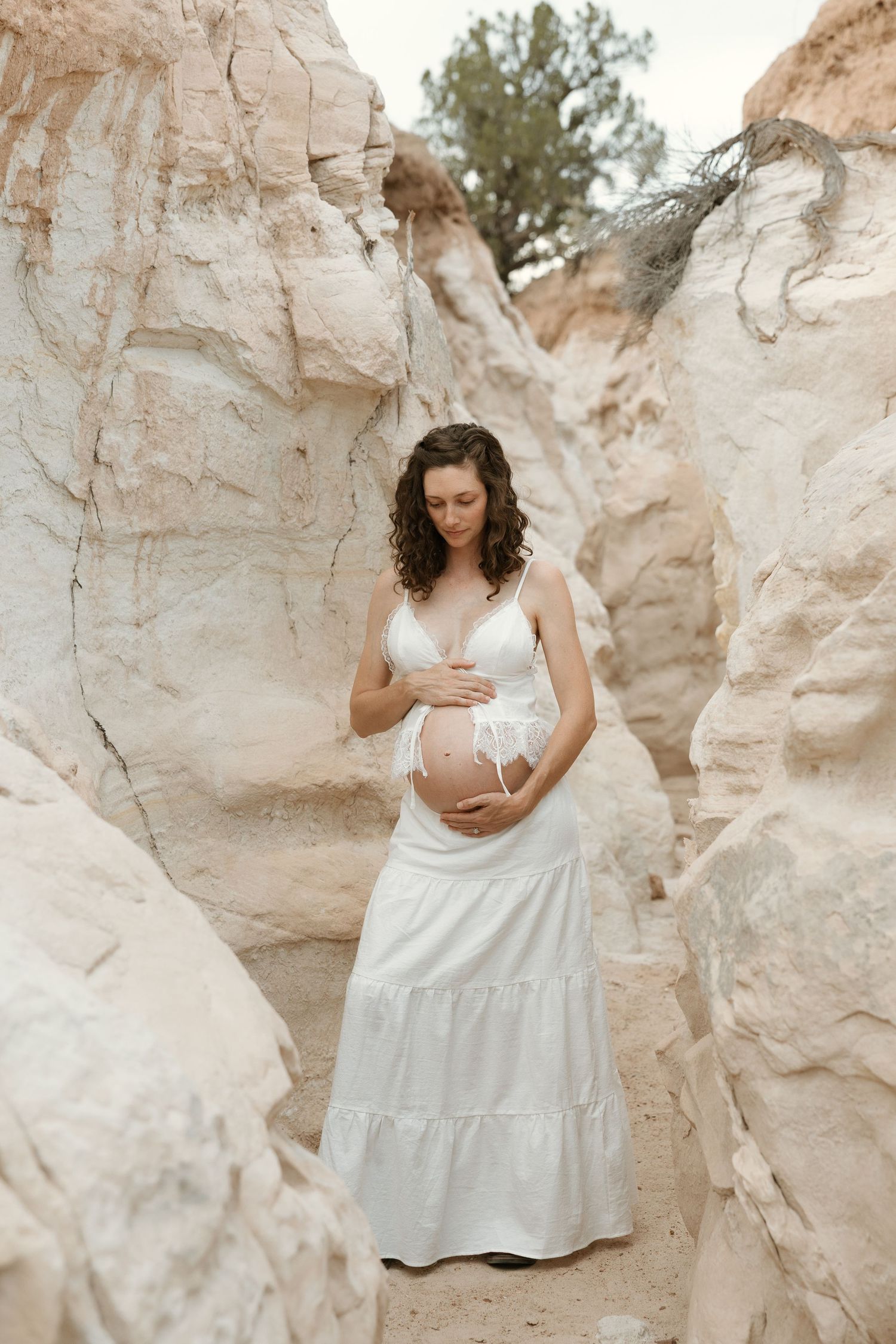 Pregnant woman in white bralette and skirt cradling baby bump among sandstone rock formations.