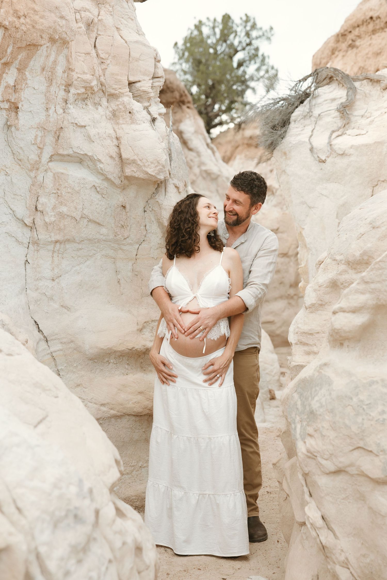 Couple embracing in sandy canyon, man holding pregnant womans belly in white maternity outfit.