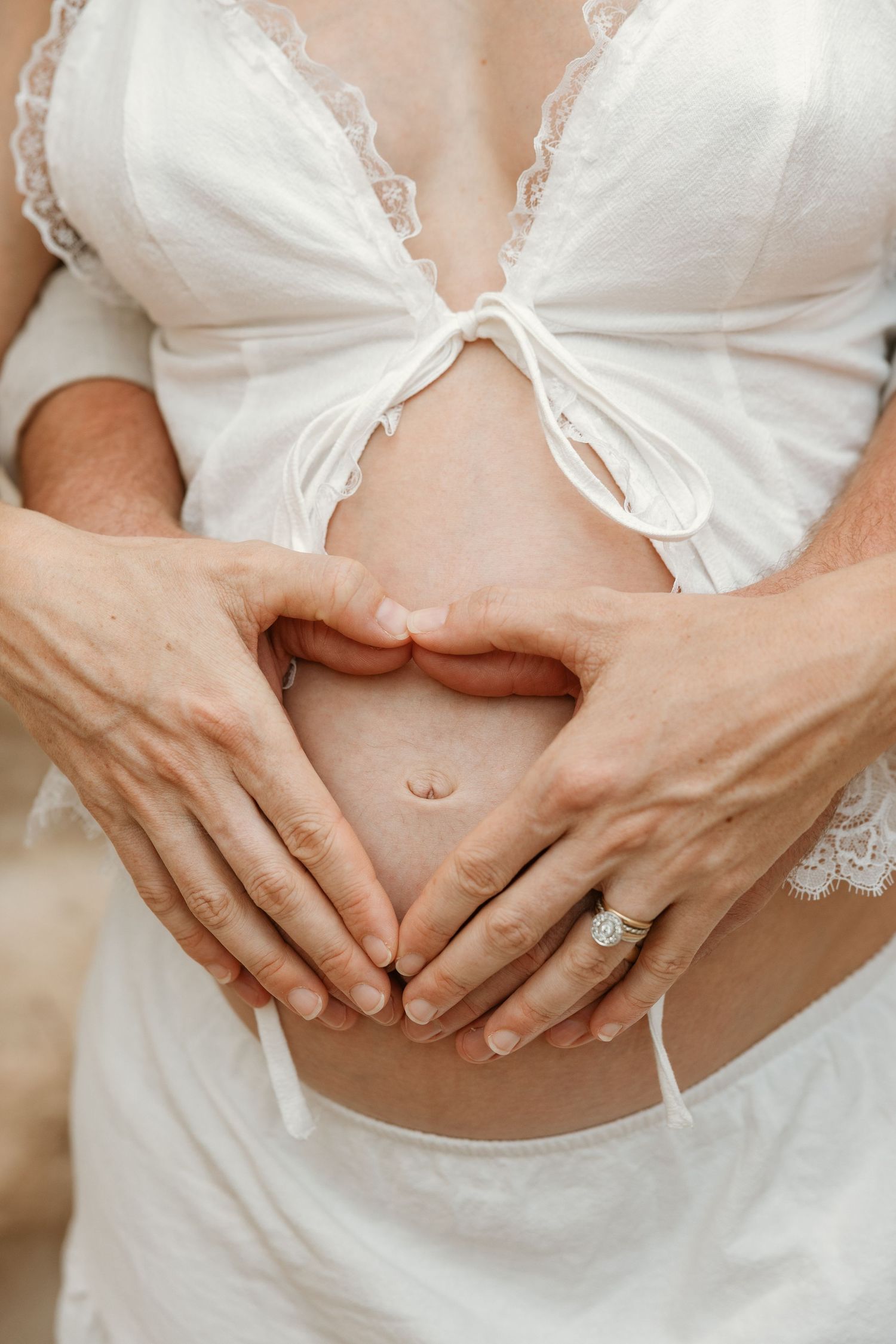 Pregnant woman in white lace top with two hands forming a heart shape on her baby bump.