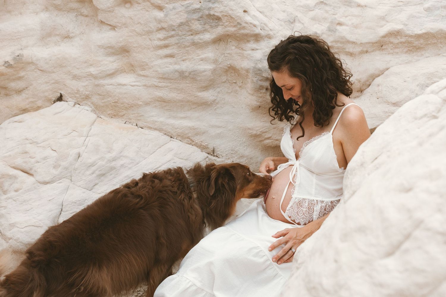Pregnant woman in white lace dress sitting against white rocks with a brown dog nuzzling her baby bump.
