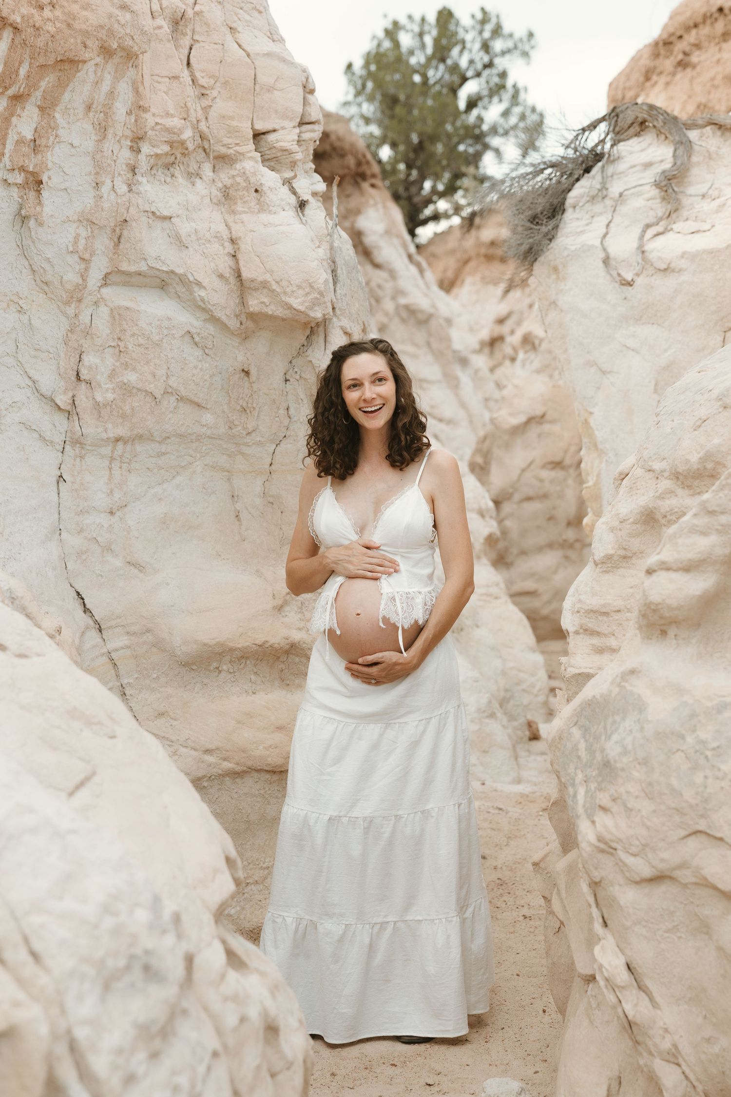 Smiling pregnant woman in white dress cradling baby bump among sandy rock formations outdoors.