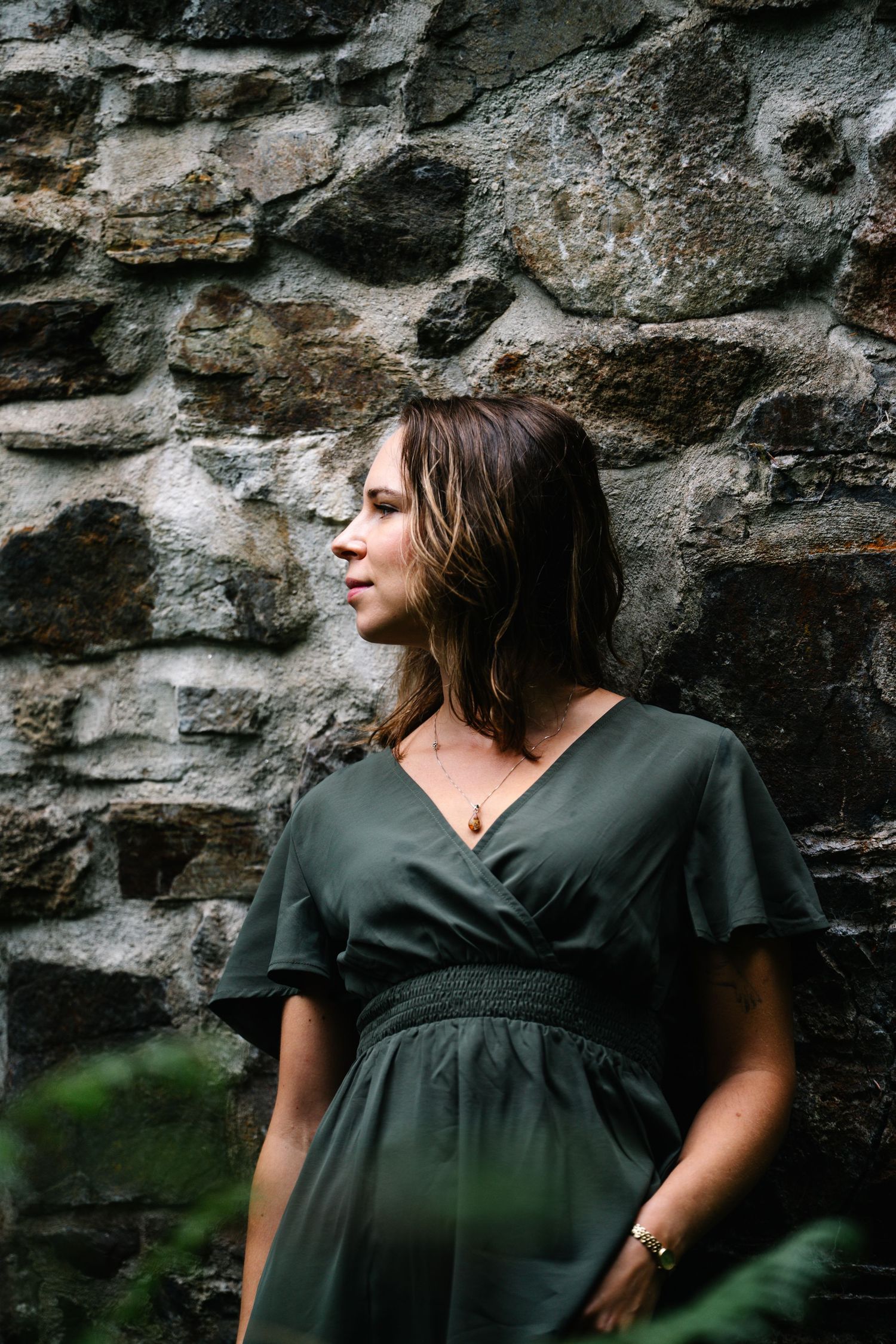 A young woman in a dark green wrap dress stands against a rustic stone wall, gazing thoughtfully to the side.
