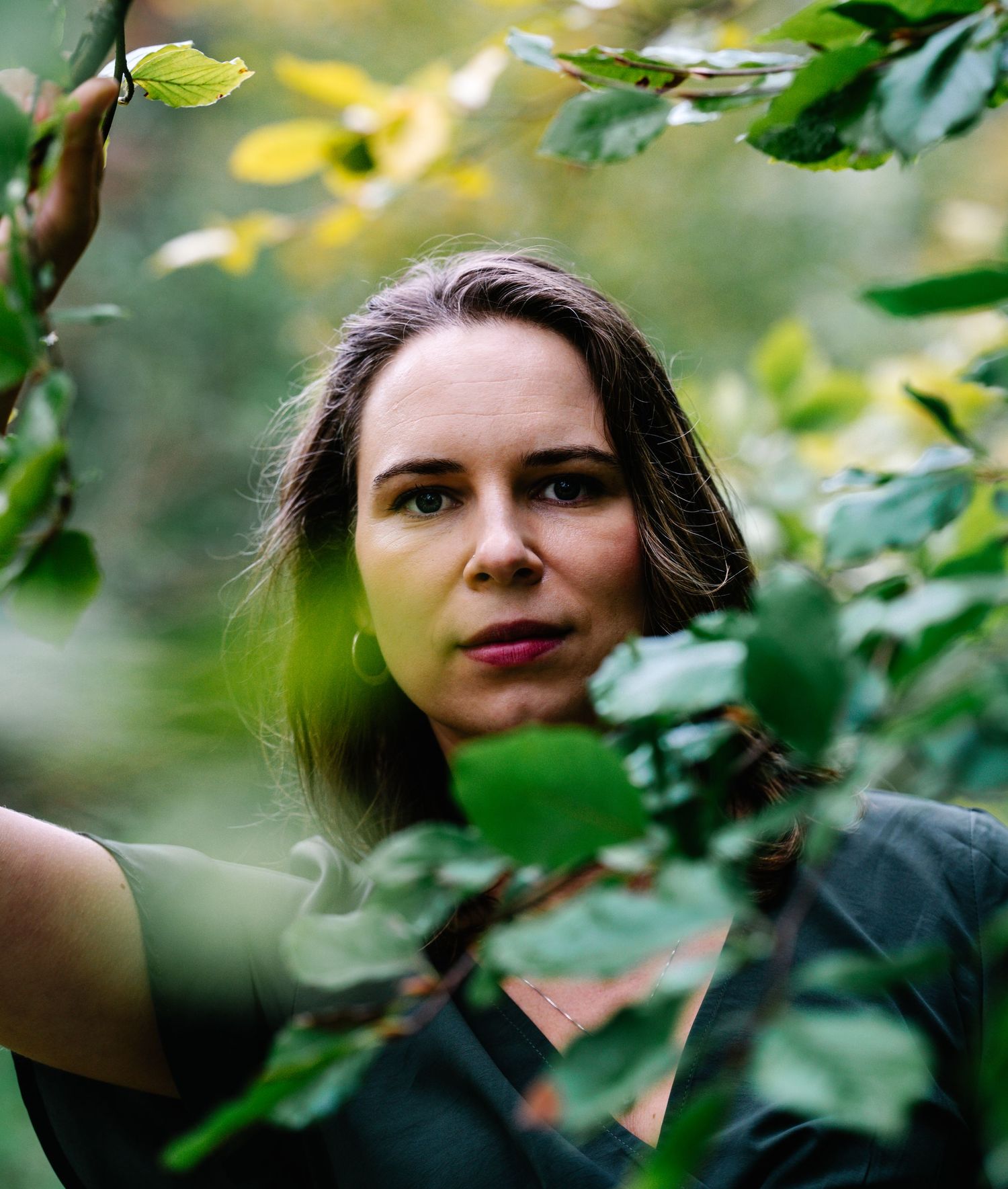 Woman peers through lush green leaves in a forest, wearing a dark top, with a serene and mysterious expression.