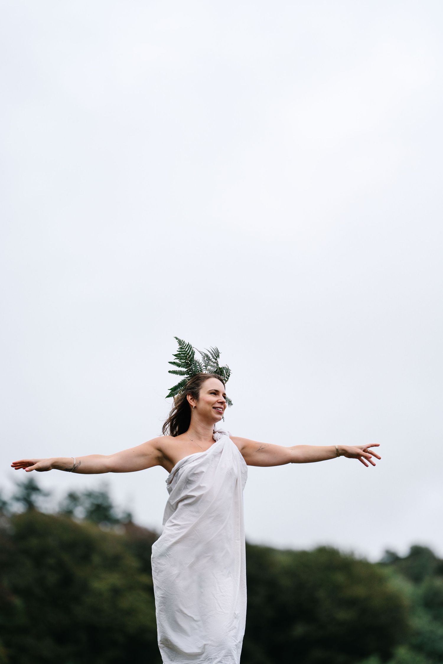 Woman in white draped dress and leaf crown stands outdoors with arms outstretched, embracing nature.