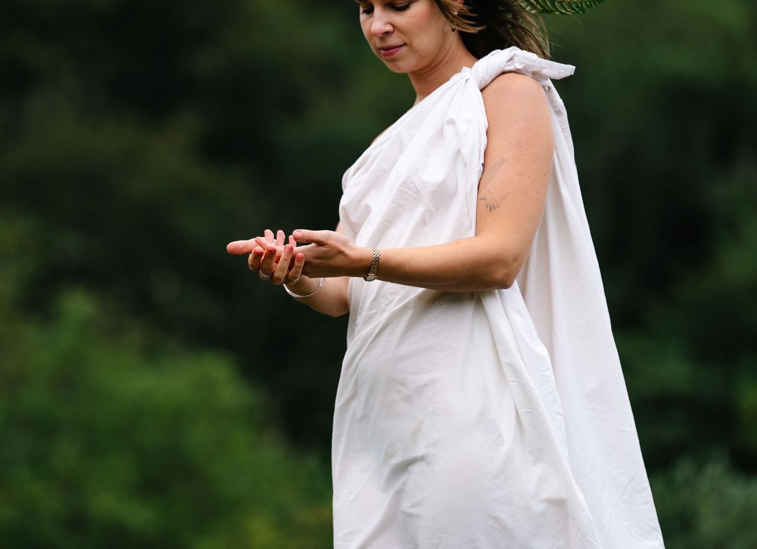 Woman in white toga dress with leaf crown gestures outdoors against green trees backdrop.