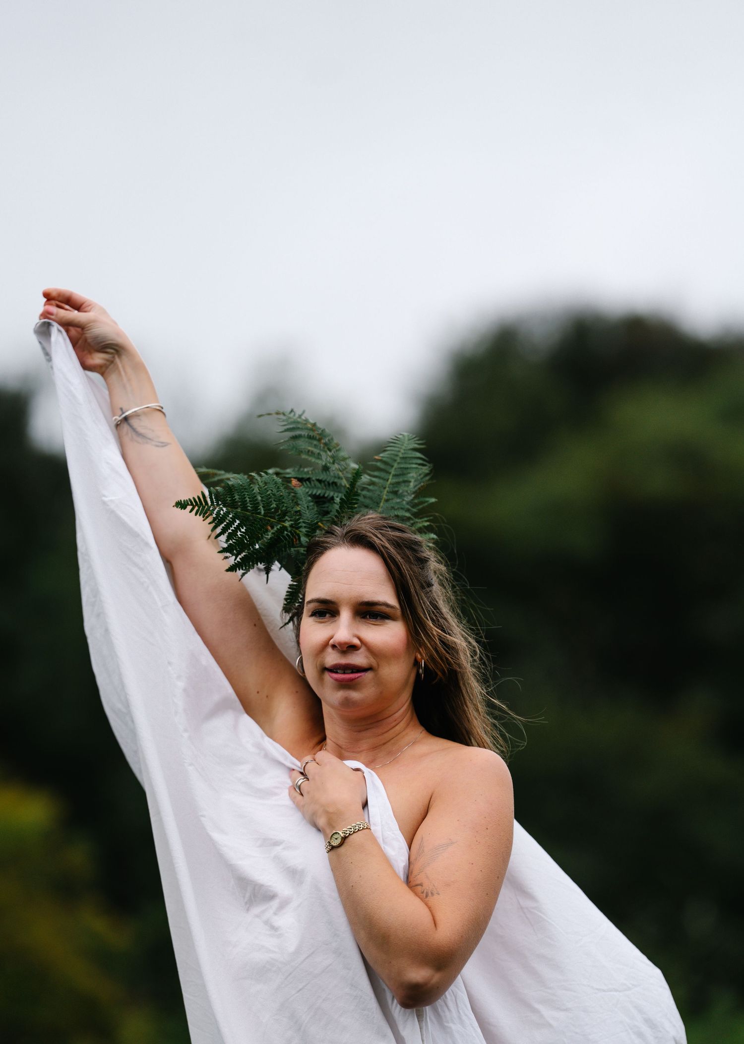 Woman holding white fabric and fern leaf outdoors with green trees in background.