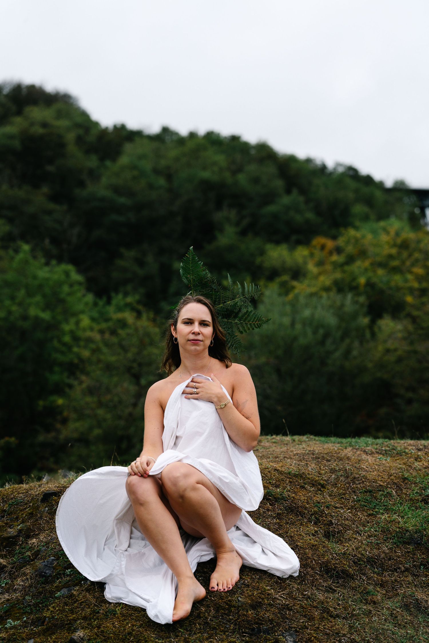 Woman in white draped fabric sits barefoot on mossy rock with fern crown, green forest background.