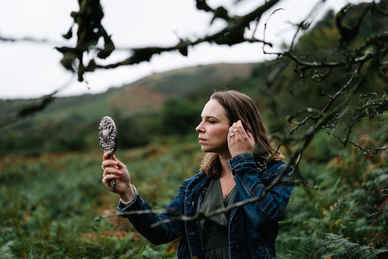 Woman in denim jacket holds ornate mirror outdoors in lush green countryside landscape.