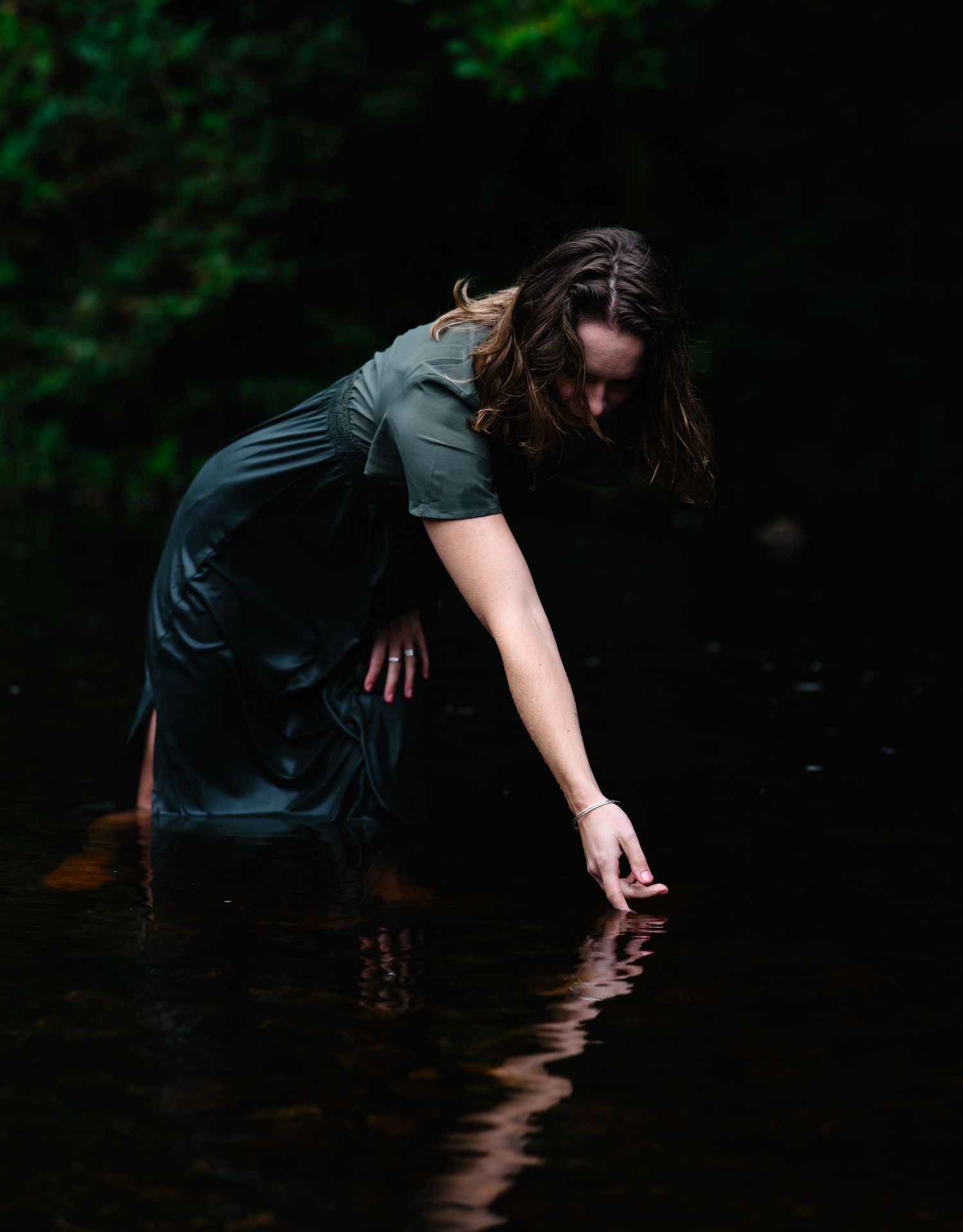 Woman in dark teal dress wading in reflective water, reaching down to touch the surface, surrounded by lush greenery.