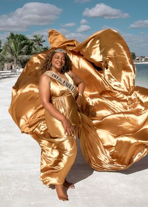 Shimmering gold gown flows dramatically in the beach breeze with palm trees and ocean in background.