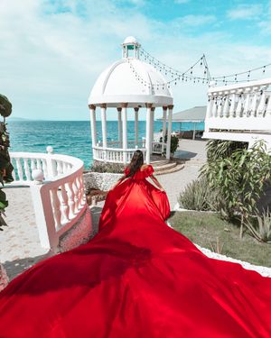 A long red dress trails dramatically leading to a white domed gazebo by the ocean.