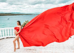 A flowing red dress billows in the wind against a coastal backdrop with white balustrades.