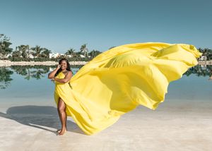 A bright yellow flowing dress catches the wind on a scenic beach with palm trees.