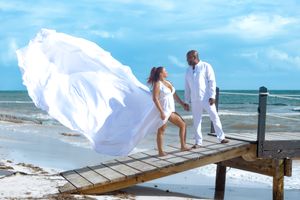 White wedding dress billows in wind as couple stands on wooden pier.